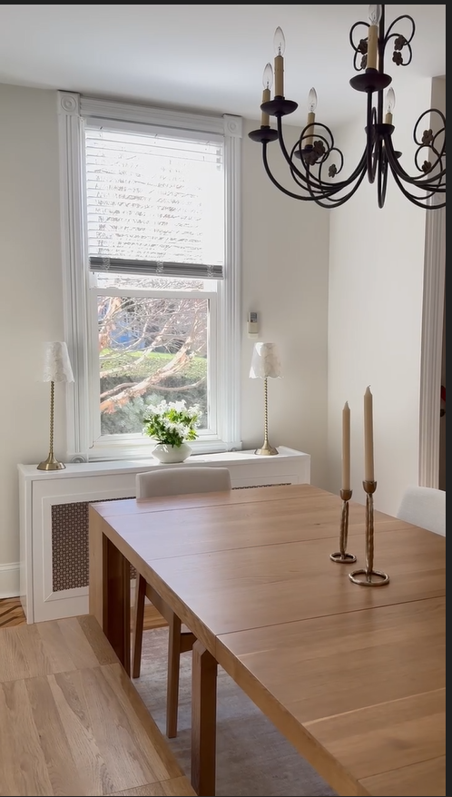 Dining room with wooden table, two candlesticks, window with blinds, two lamps, chandelier, and a potted plant on the window sill.