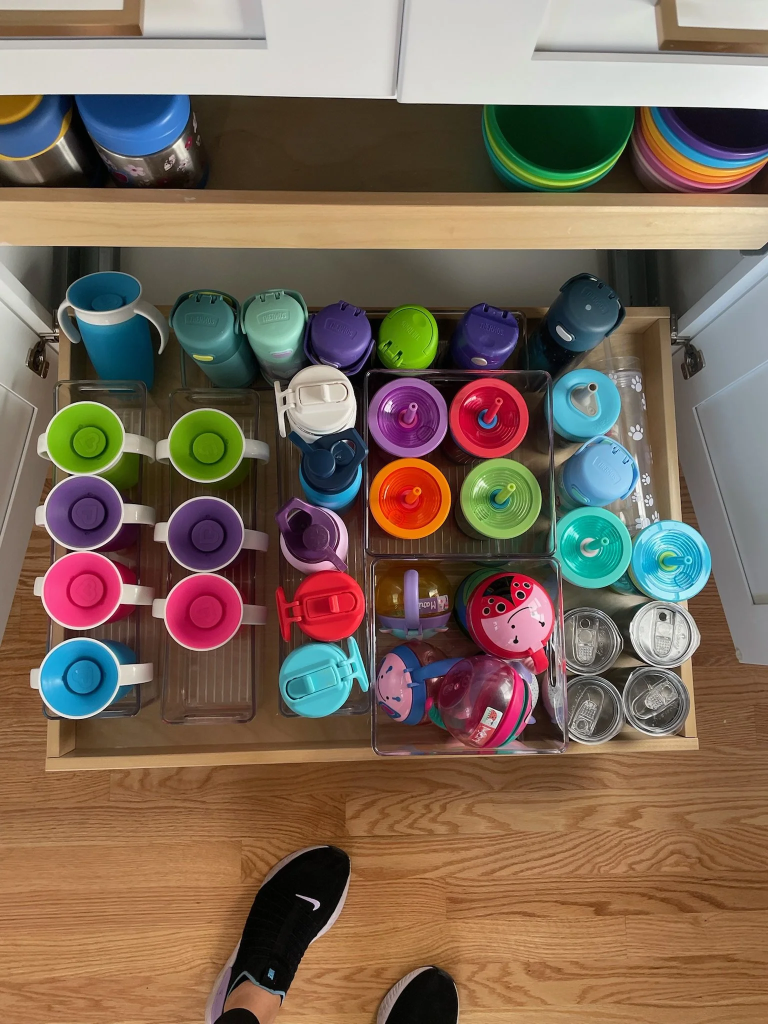 Open kitchen drawer filled with colorful drink bottles, cups, and lids, with a person's feet visible at the bottom.