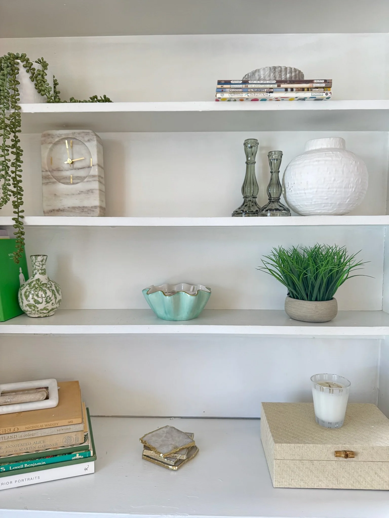 White bookshelf with decorative items including books, a clock, glass candle holders, a white textured vase, a green plant, and small coaster tiles.