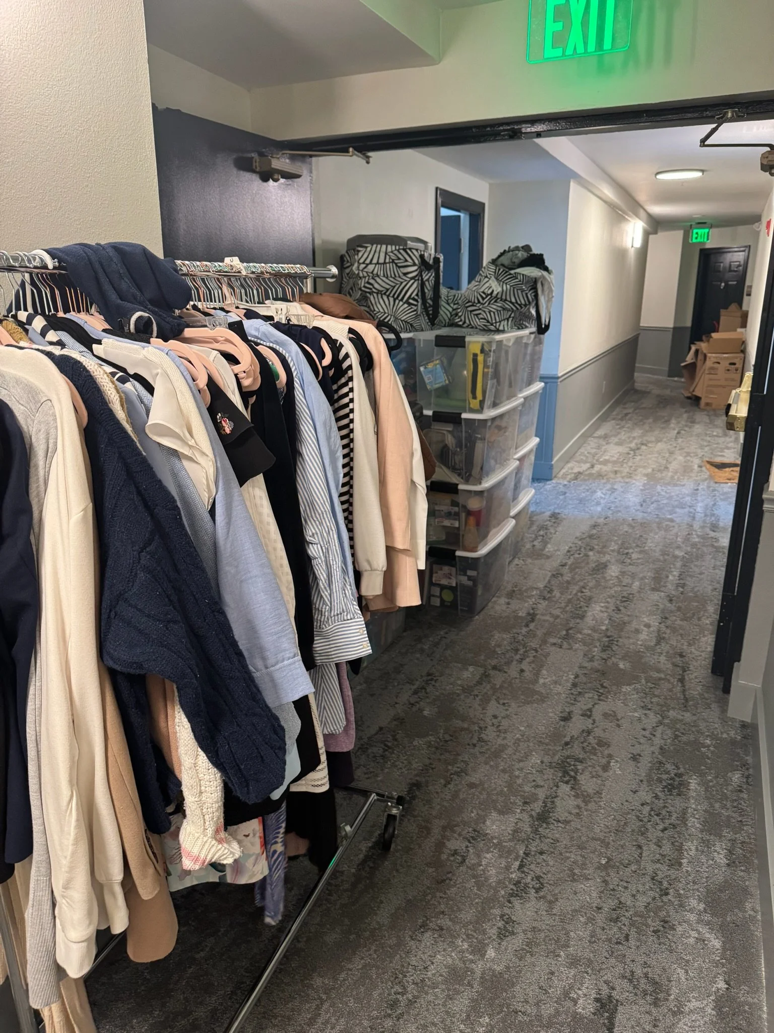 Clothes hanging on a rack next to plastic storage containers in a hallway with an exit sign above.