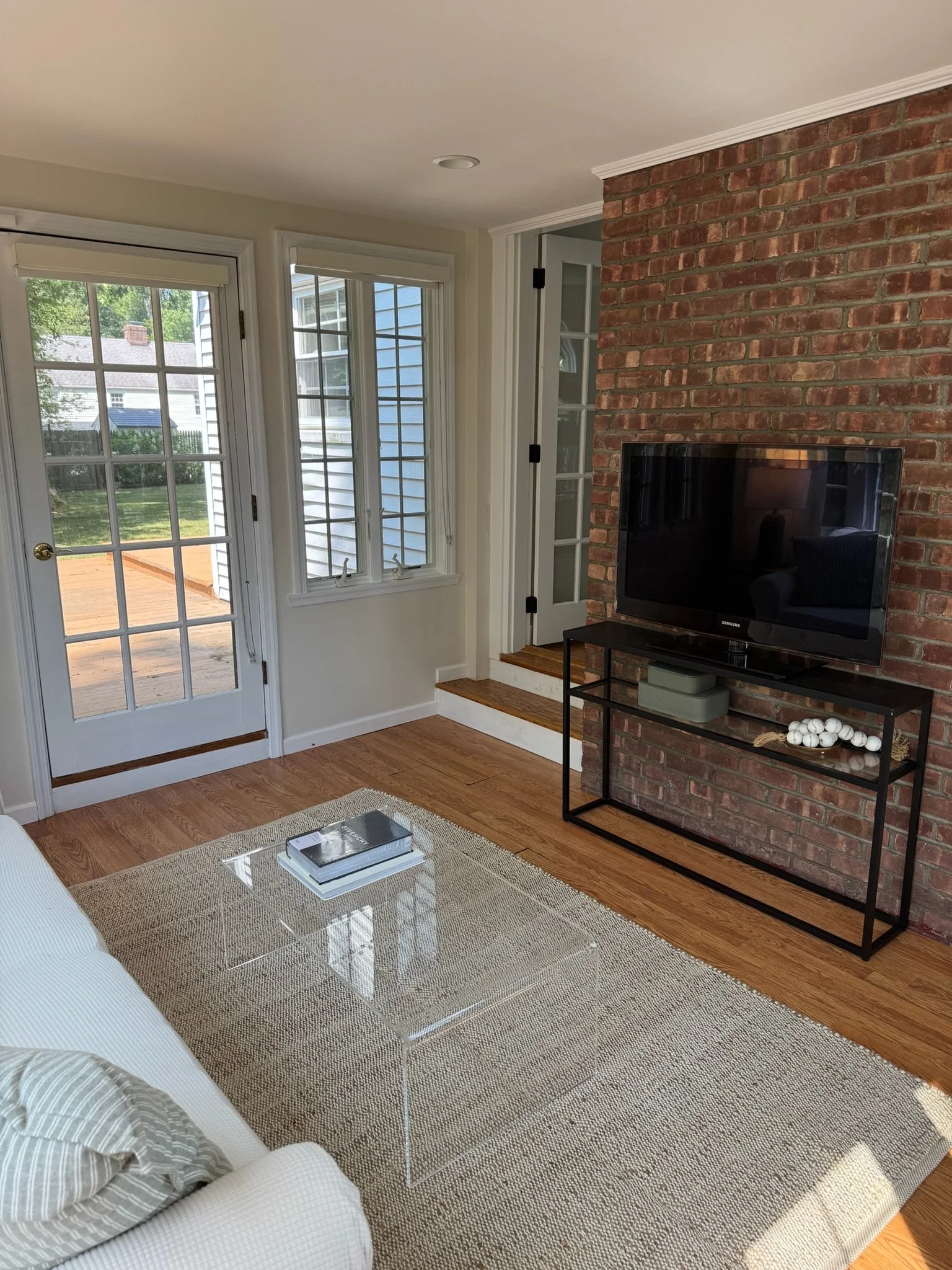 Cozy living room with a brick accent wall, mounted flat-screen TV, glass coffee table, light-colored sofa, wooden floors, and large windows with a door leading outside to a wooden deck.