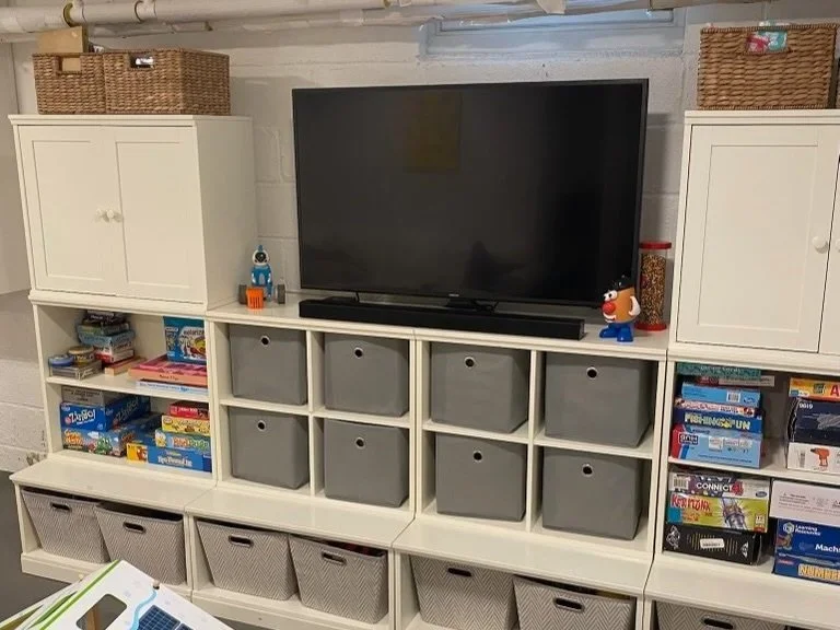 A white entertainment center with a flat-screen TV on top, various board games and toys on the shelves, and storage bins in the cubbies.