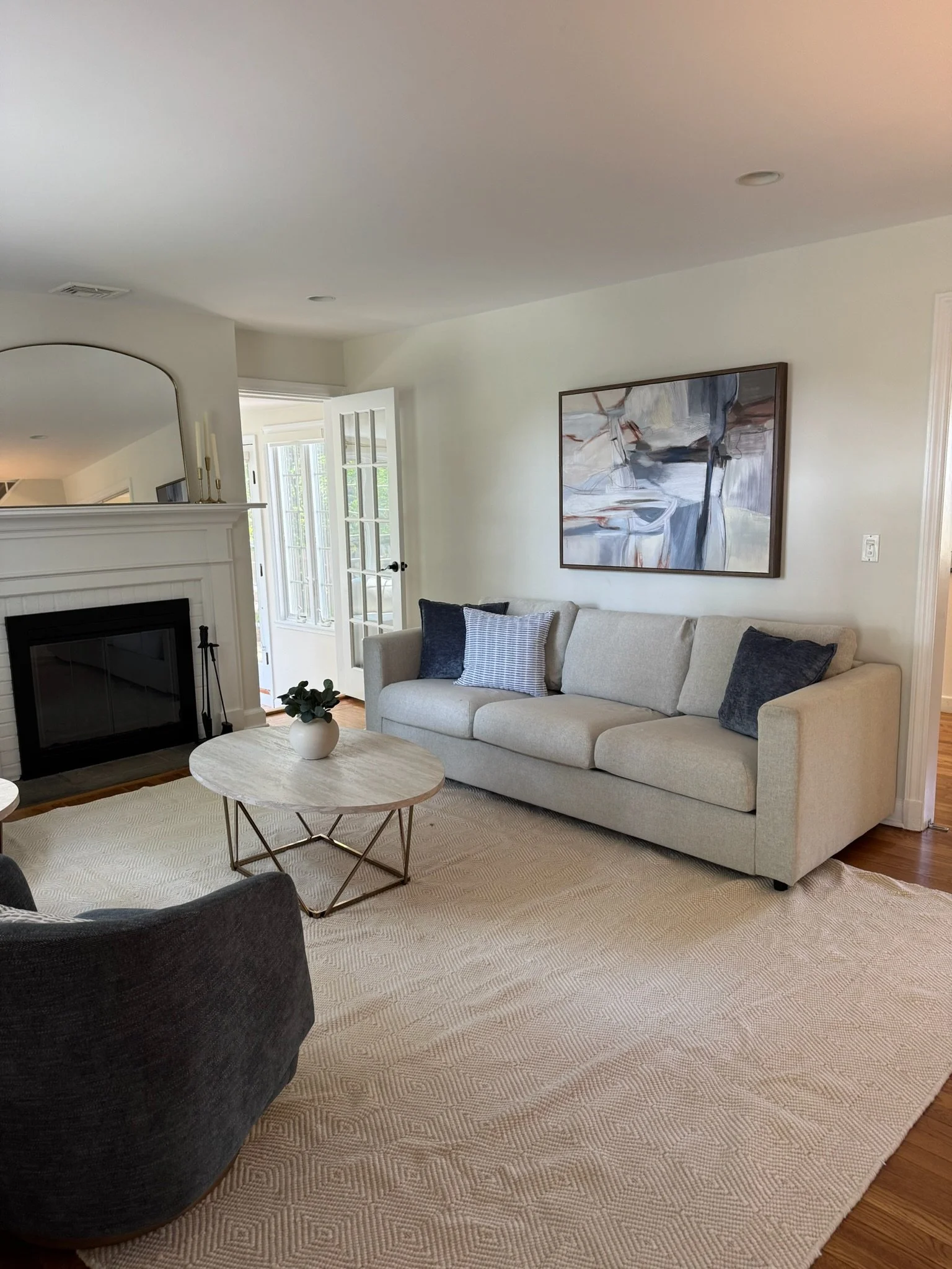 Living room with white sofa, abstract art on wall, beige rug, black armchair, white marble coffee table with plant, fireplace, open door with windows.