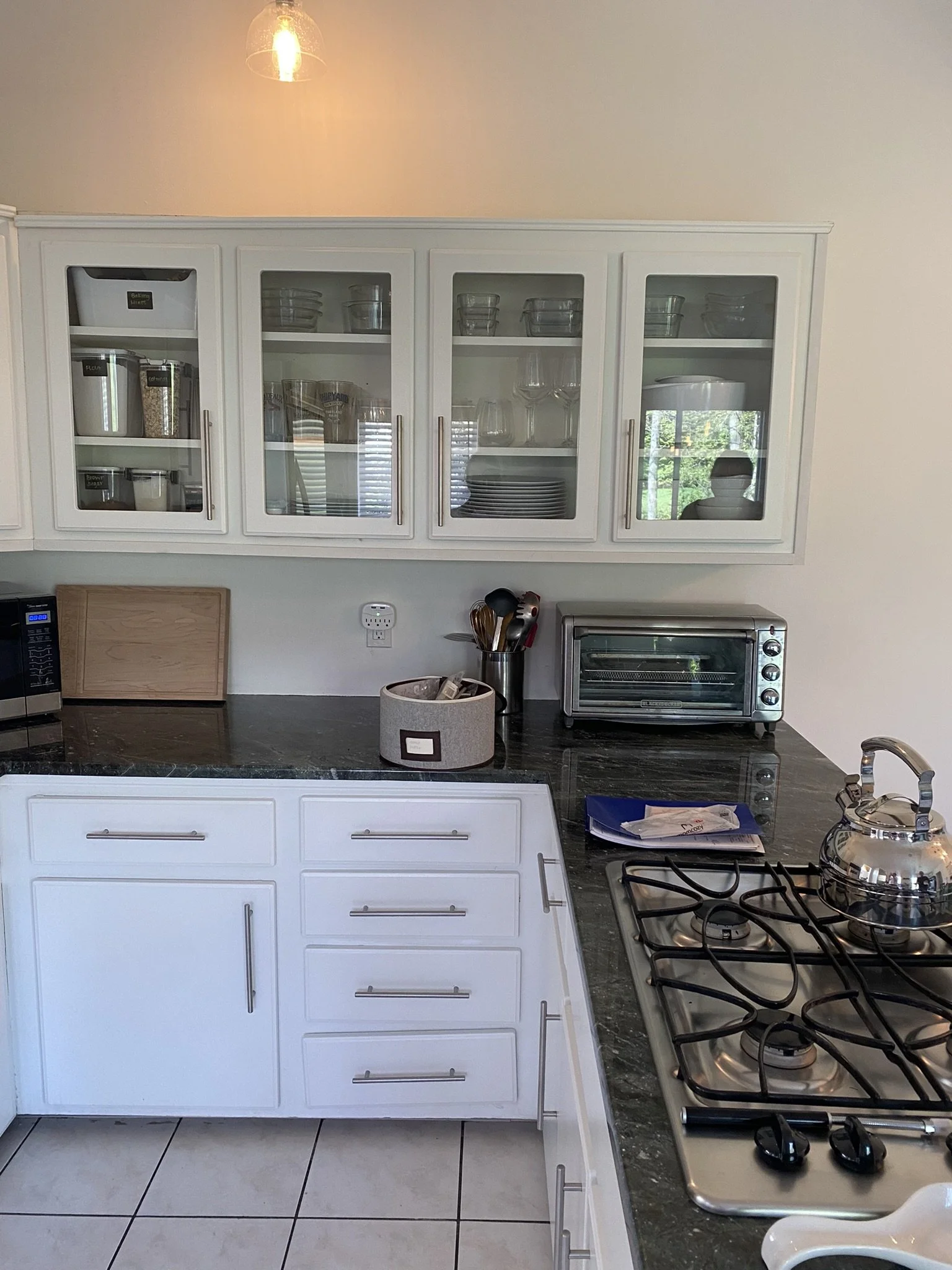 White kitchen cabinets with glass doors, black granite countertop, stainless steel stovetop with kettle, toaster oven, and various kitchen utensils in a container, with a tiled floor.