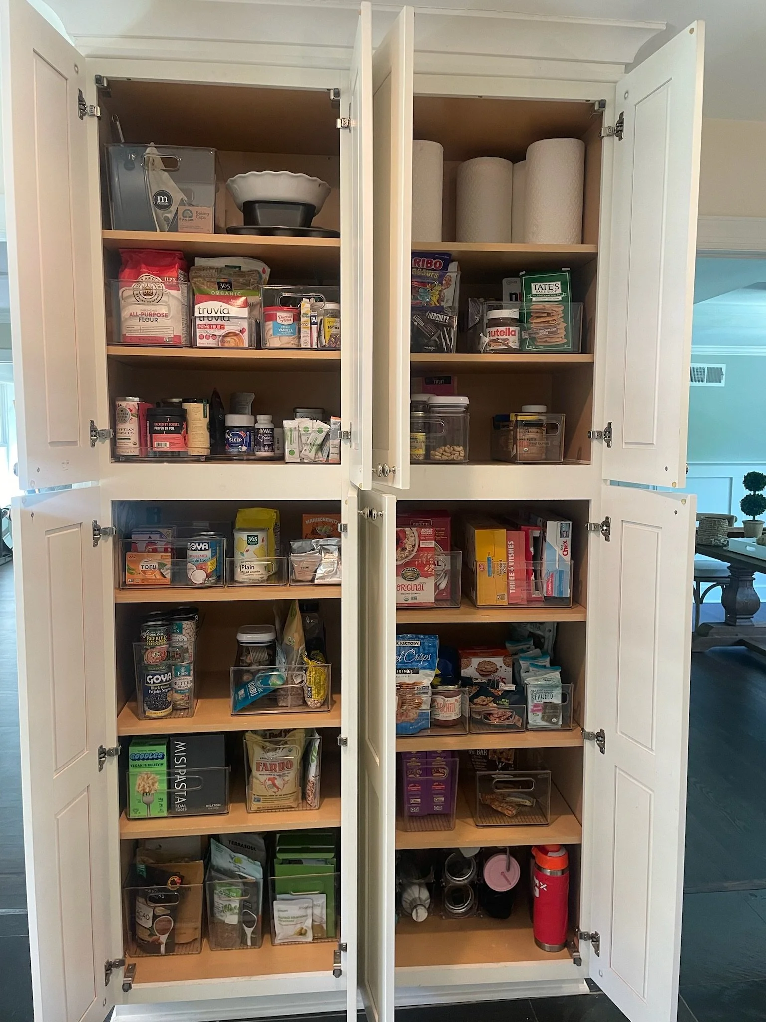 Open kitchen pantry cabinet with four shelves filled with food items, snacks, and kitchen supplies.