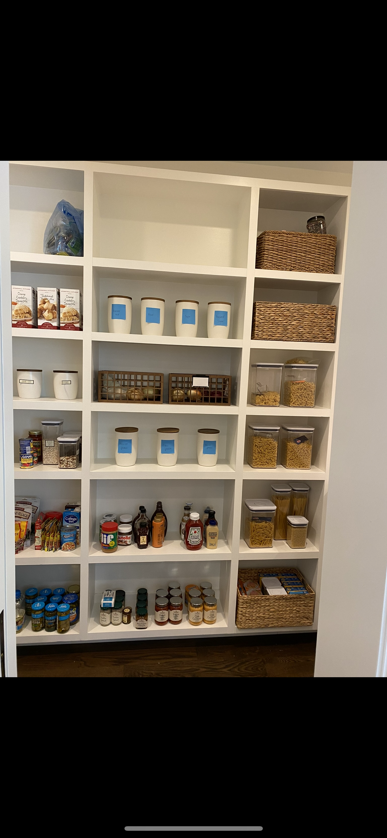 Pantry with various food and condiment containers, jars, boxes, and baskets organized on white shelves.
