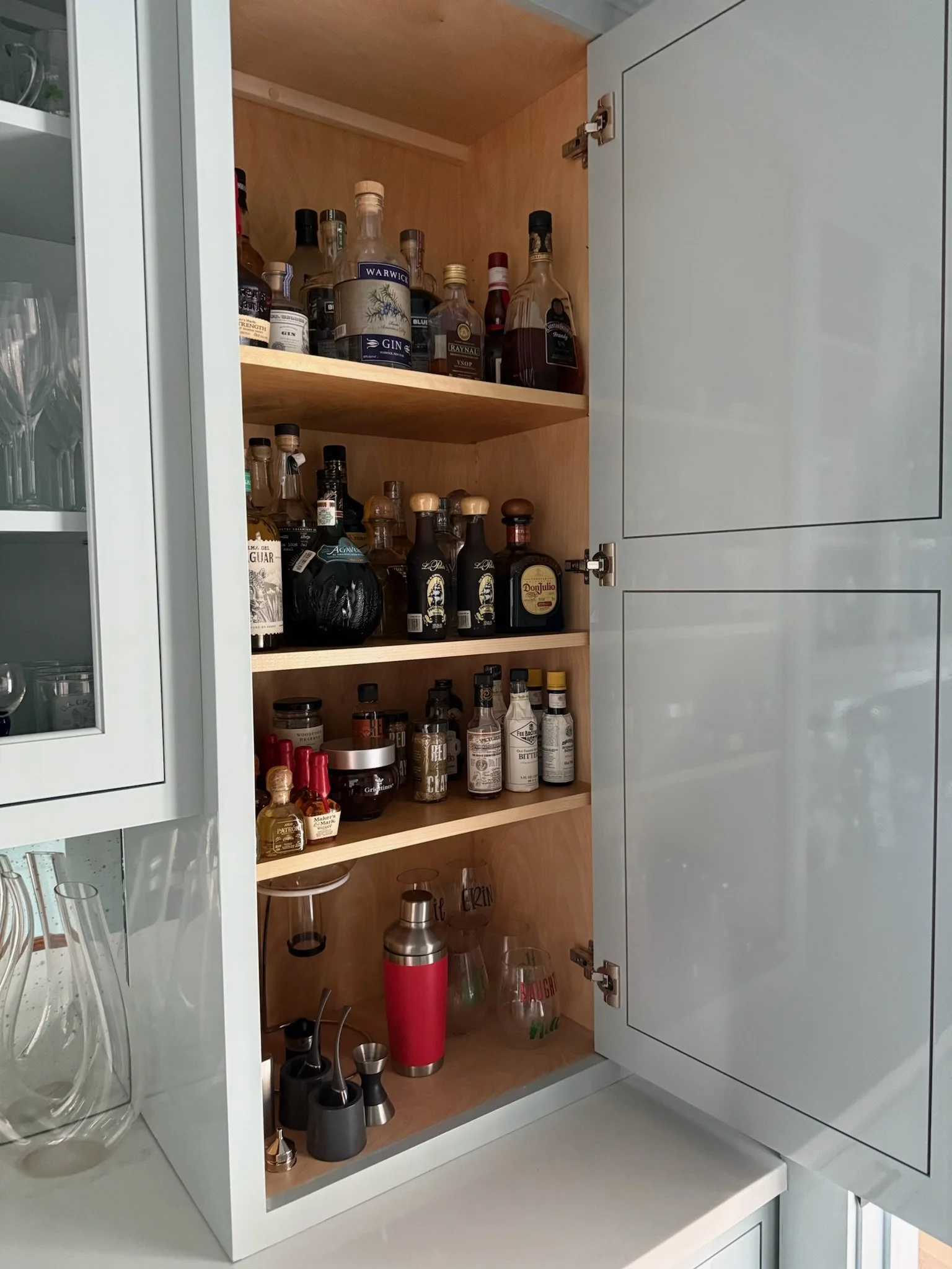 Open kitchen cabinet with shelves displaying various bottles of alcohol, mixers, and barware.