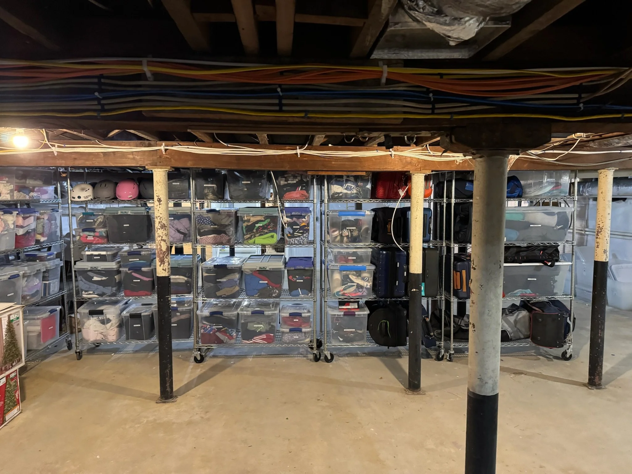 Storage area with metal shelves filled with transparent plastic bins containing clothing, and some lockers or suitcases. Helmets are on the top shelf, and the space is supported by metal poles with a wooden ceiling above.