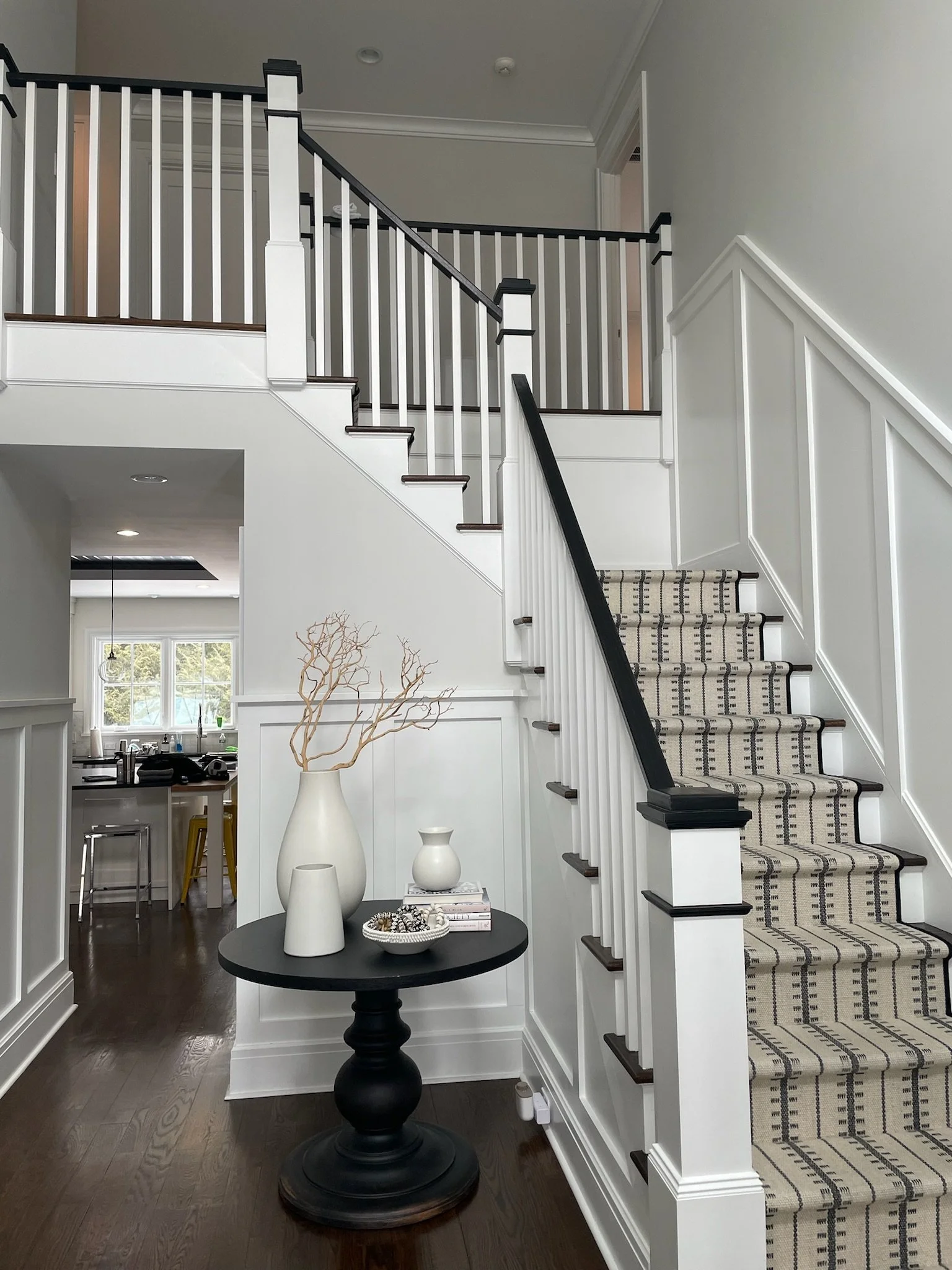 Interior of a home featuring a staircase with a black handrail and patterned carpet, a black round table with decorative white vases, and a view into the kitchen area with a window and barstools.