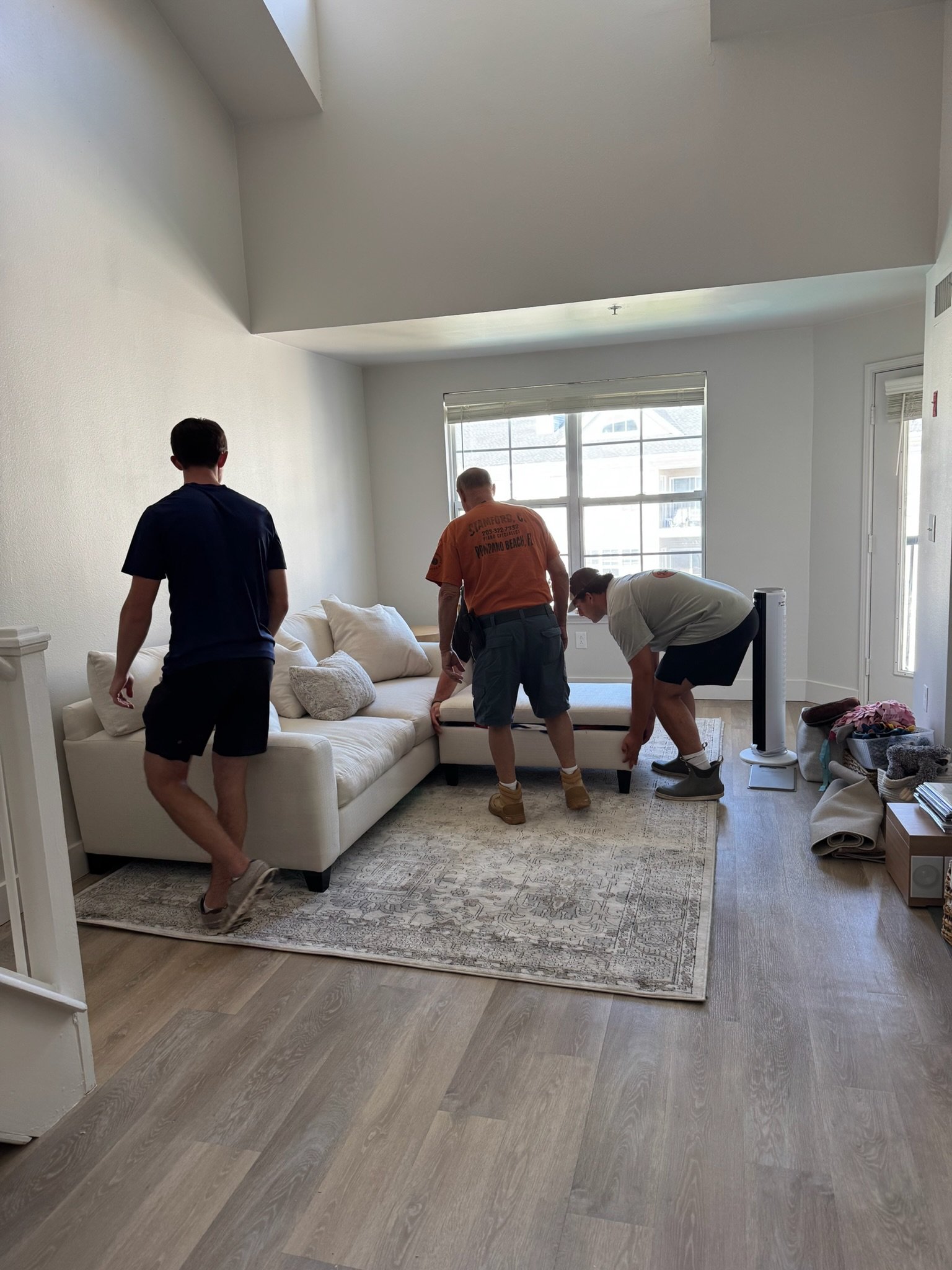 Three men are moving a piece of furniture in a living room with a white sofa, rug, and window, working together to position it.