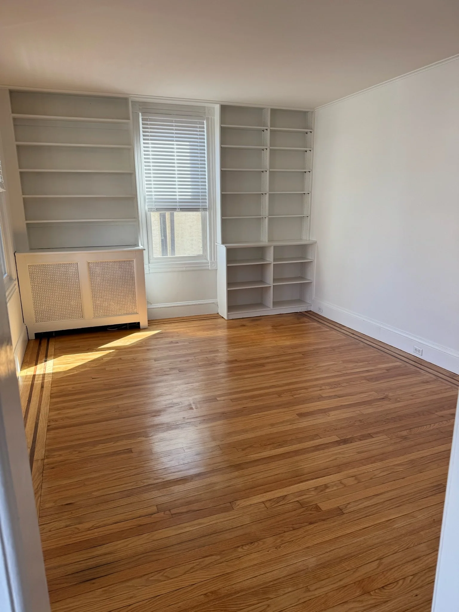Empty room with hardwood floors, white walls, and built-in white bookshelves near a window with blinds.