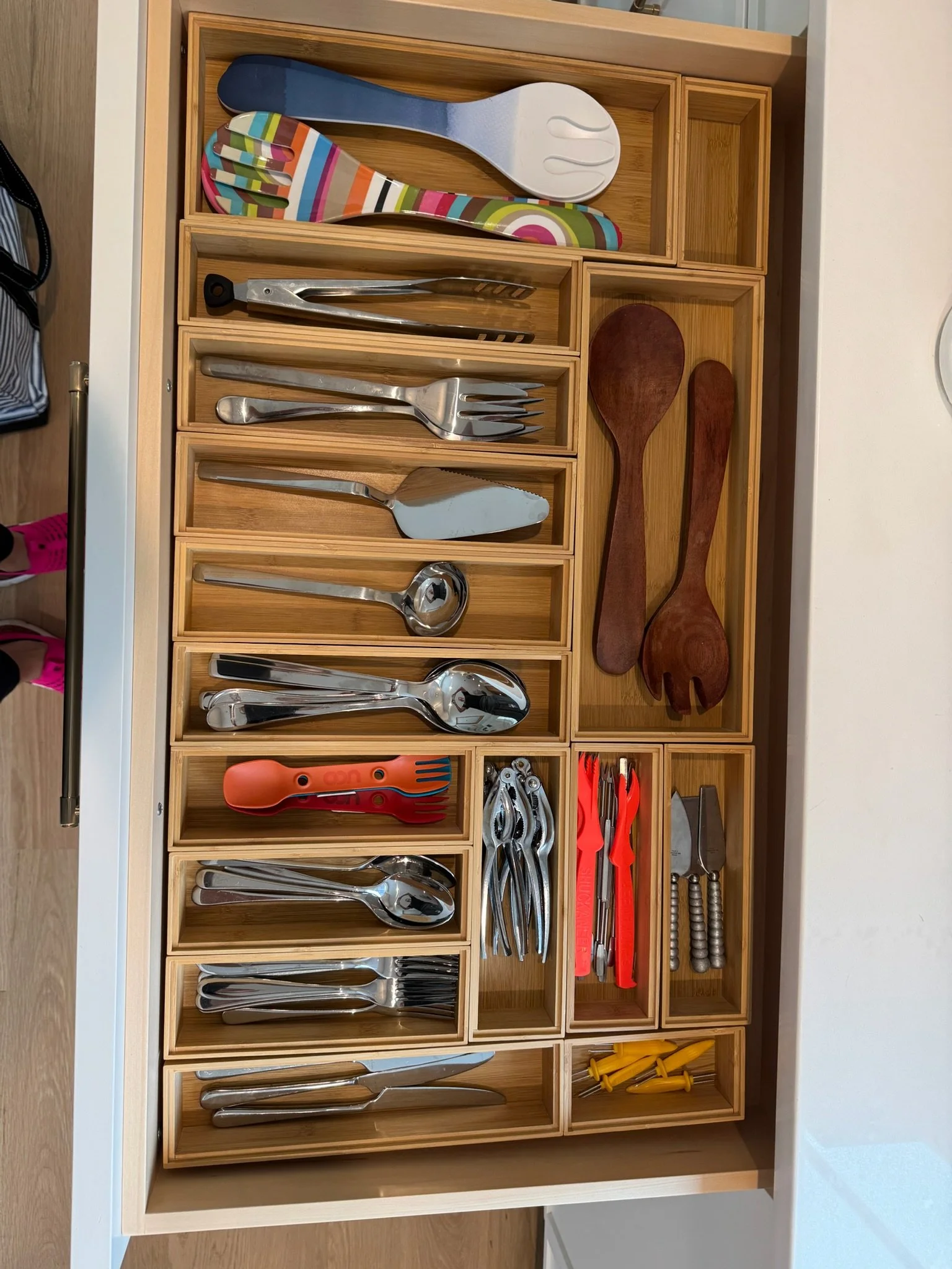 A wooden drawer organizer with various kitchen utensils including spatulas, tongs, spoons, forks, knives, pie server, and colorful toddler utensils.