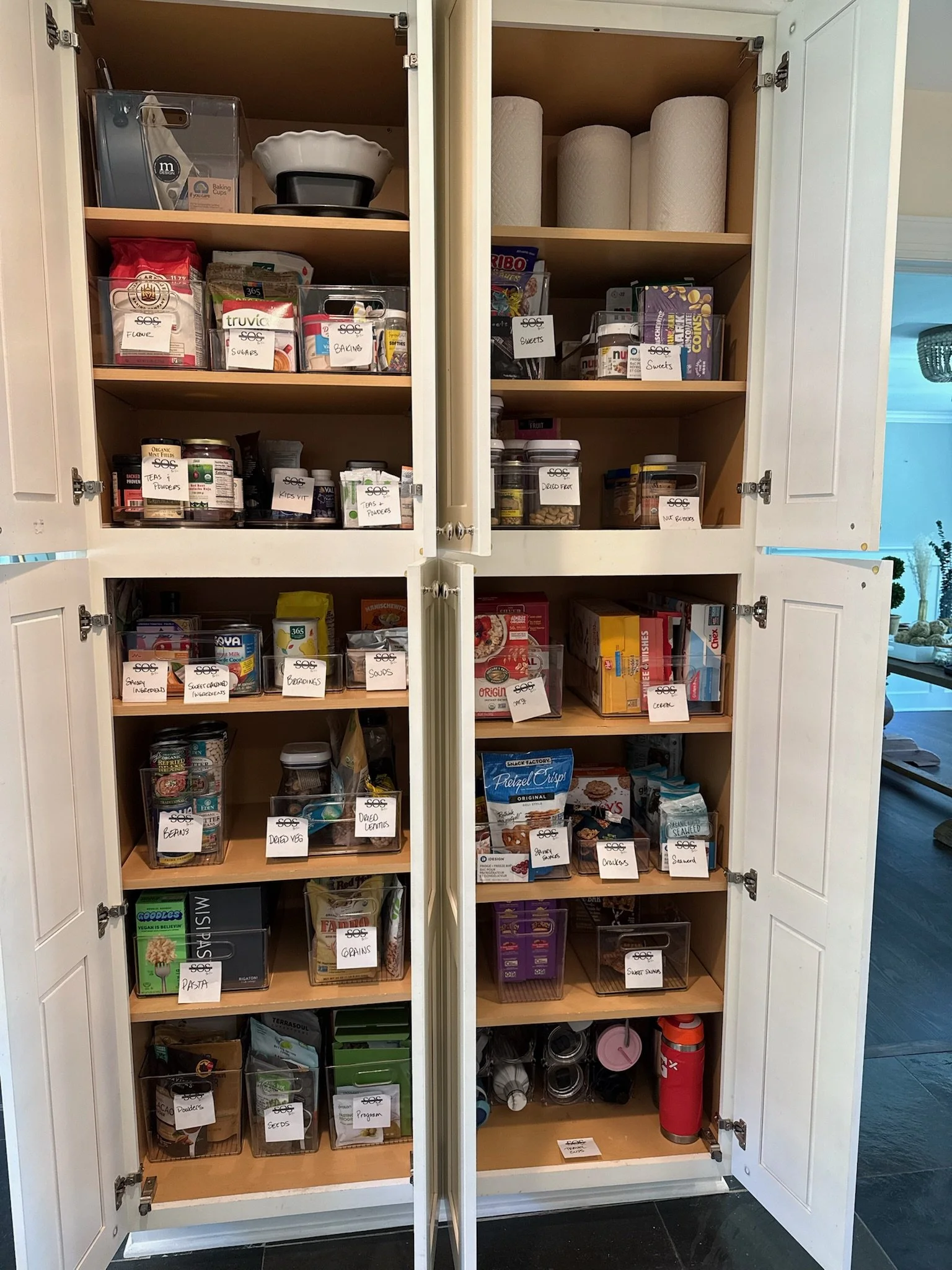 A pantry cupboard with four shelves filled with labeled containers of various food items, paper towels on the top shelf, and some storage bins on the bottom shelf.