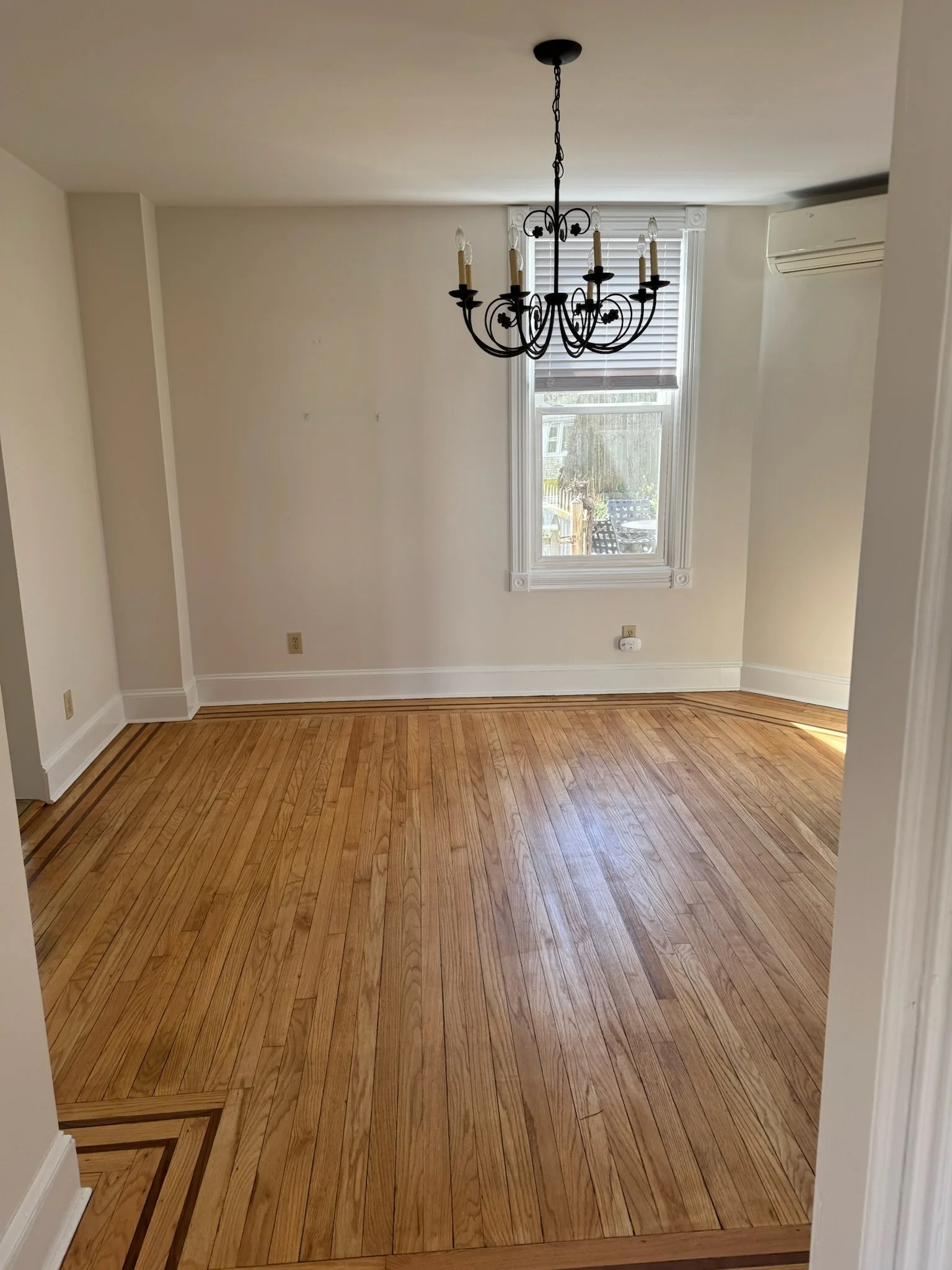 Empty room with hardwood floor, white walls, a window with blinds, a chandelier hanging from the ceiling, and an air conditioning unit.