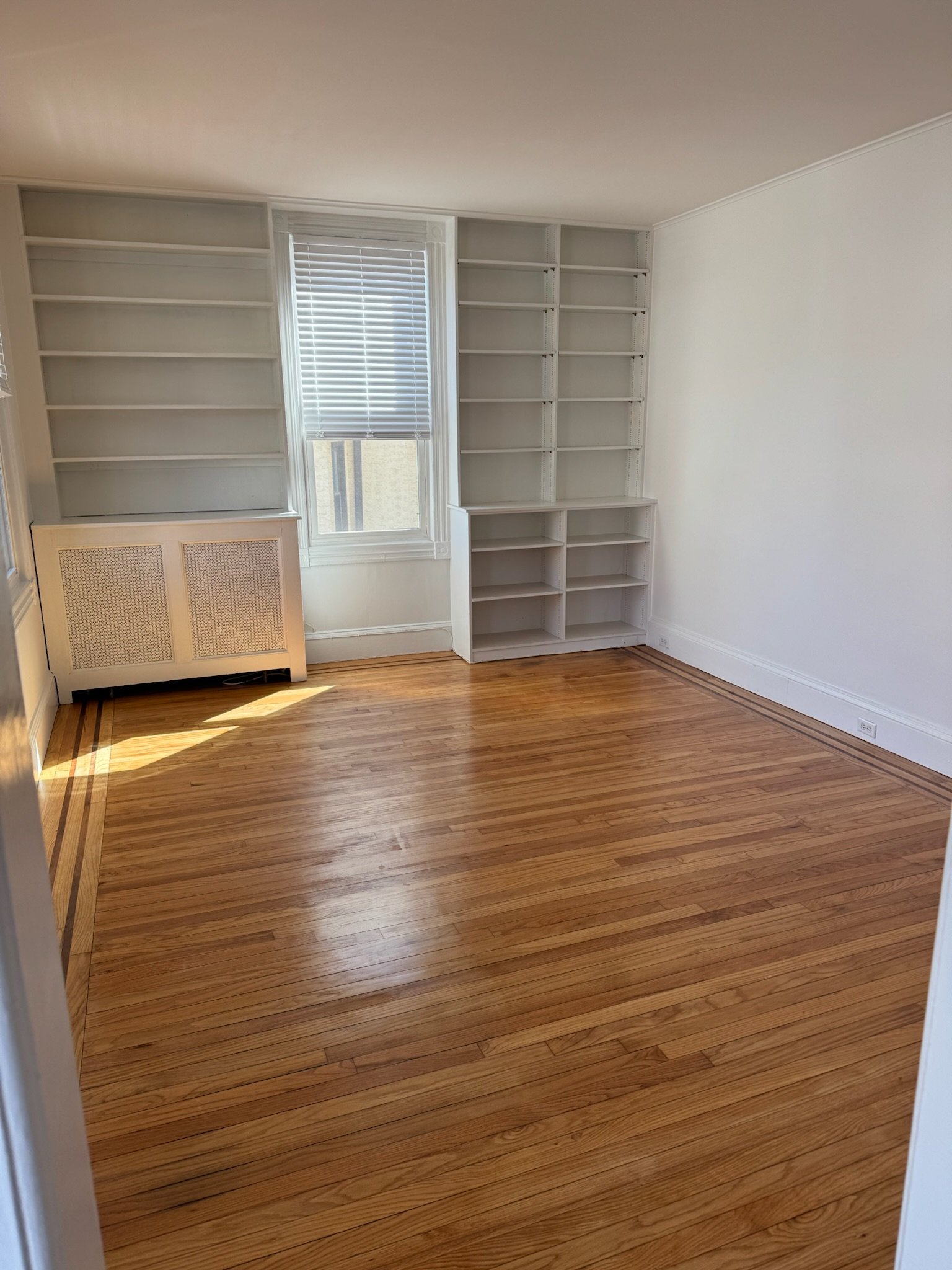 Empty room with wooden floors, white walls, built-in white bookshelves, and a window with blinds, allowing sunlight to fill the room.