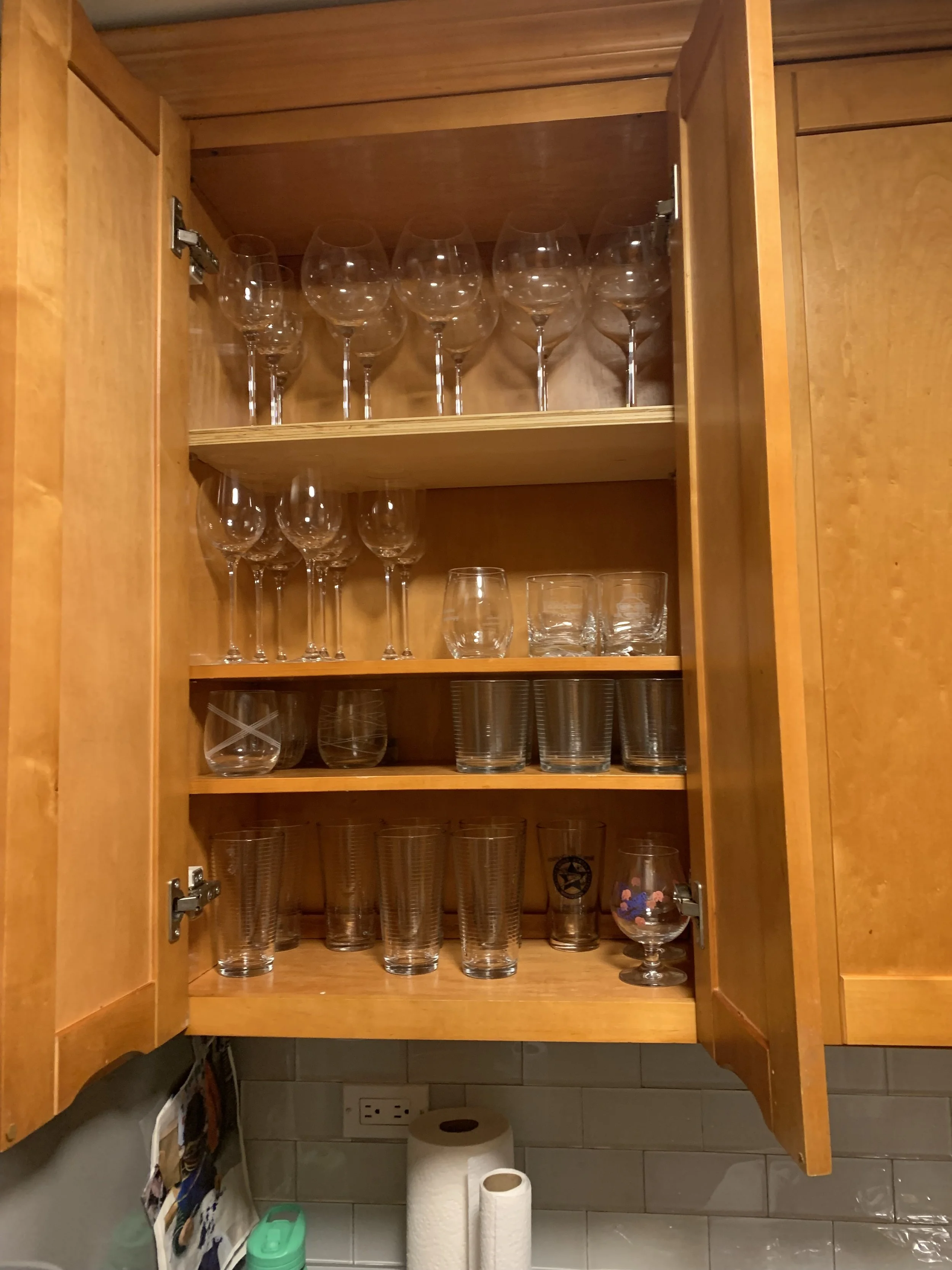 Open wooden kitchen cabinet with various glasses including wine glasses, tumblers, and compotes on shelves.