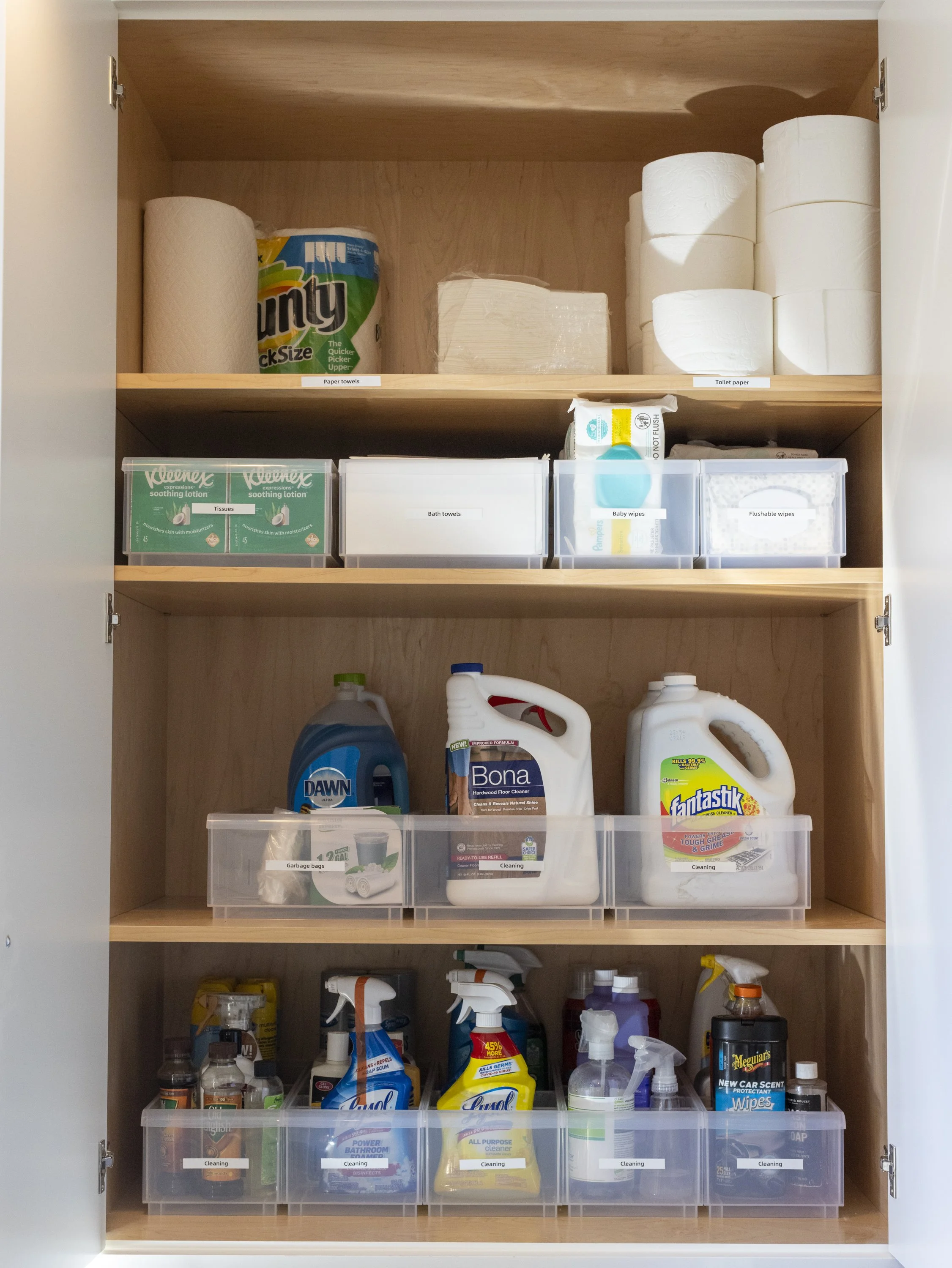 A cabinet with three shelves filled with cleaning supplies, paper towels, toilet paper, tissues, bath towels, baby wipes, and garbage bags.