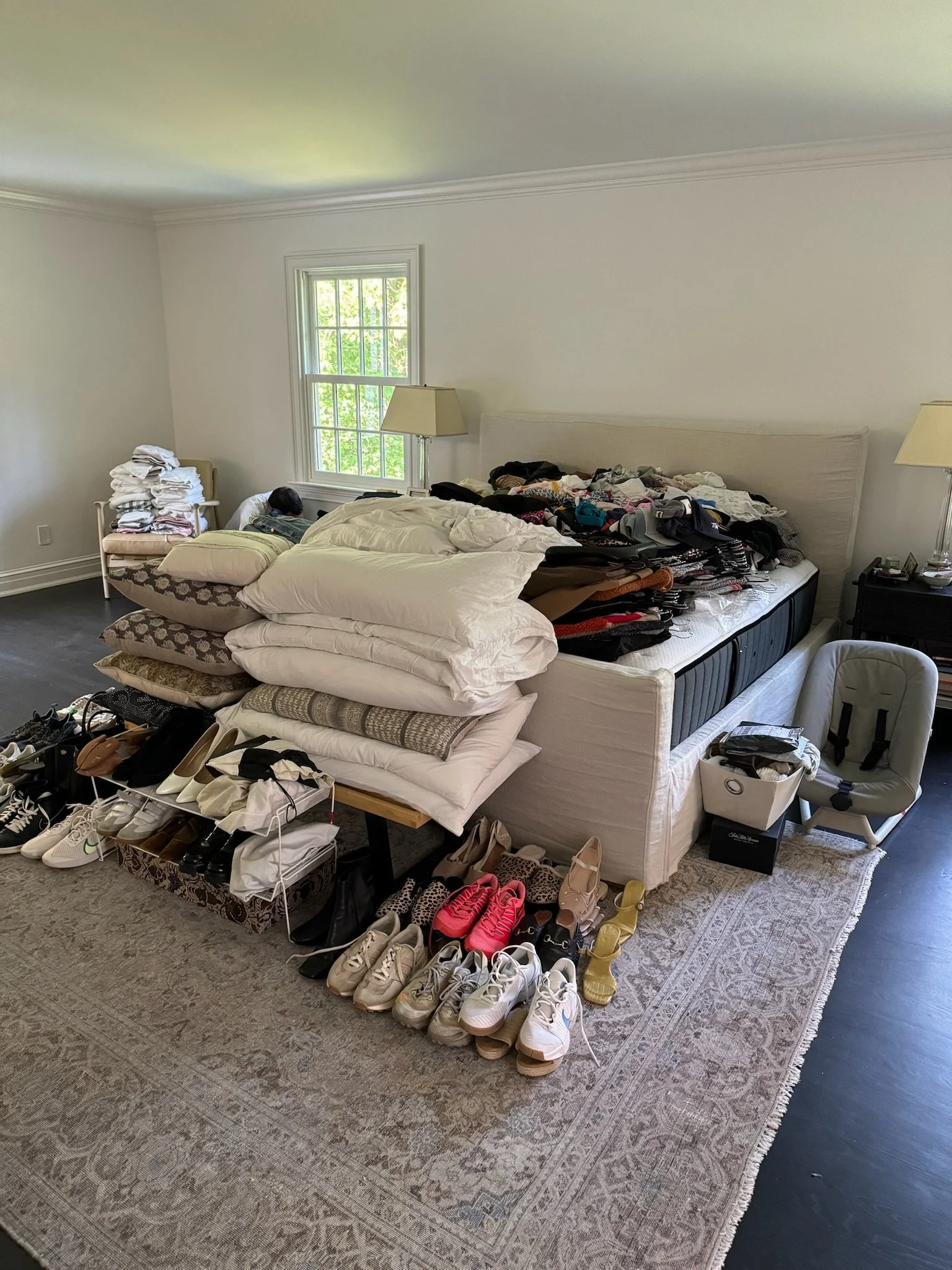 A bedroom with a large bed covered in clothes, shoes, and pillows. Shoes are lined up on the floor in front of the bed. There are nightstands with lamps on either side of the bed and a window with green trees outside.