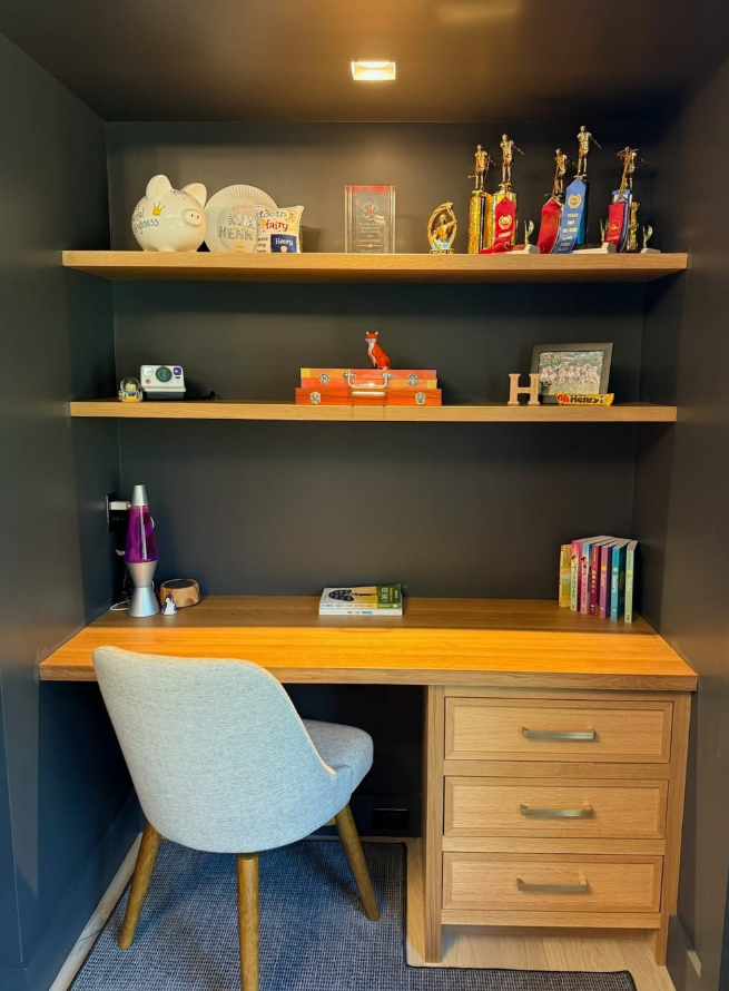 A small workspace with a wooden desk, a grey upholstered chair, and dark grey walls. The desk has books, a purple lava lamp, and a small lamp. Two wooden shelves above hold trophies, awards, and decorative items, including a fox figurine and framed photos.