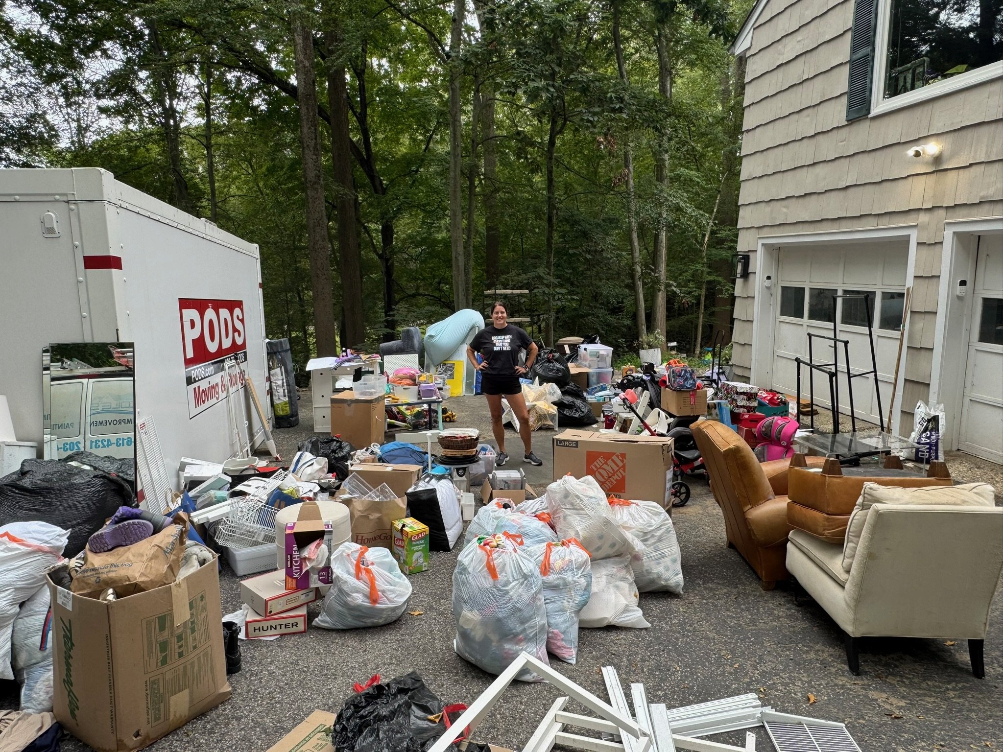 Person standing outside among various household items, bags, and boxes in a driveway next to a house with a wooded background.