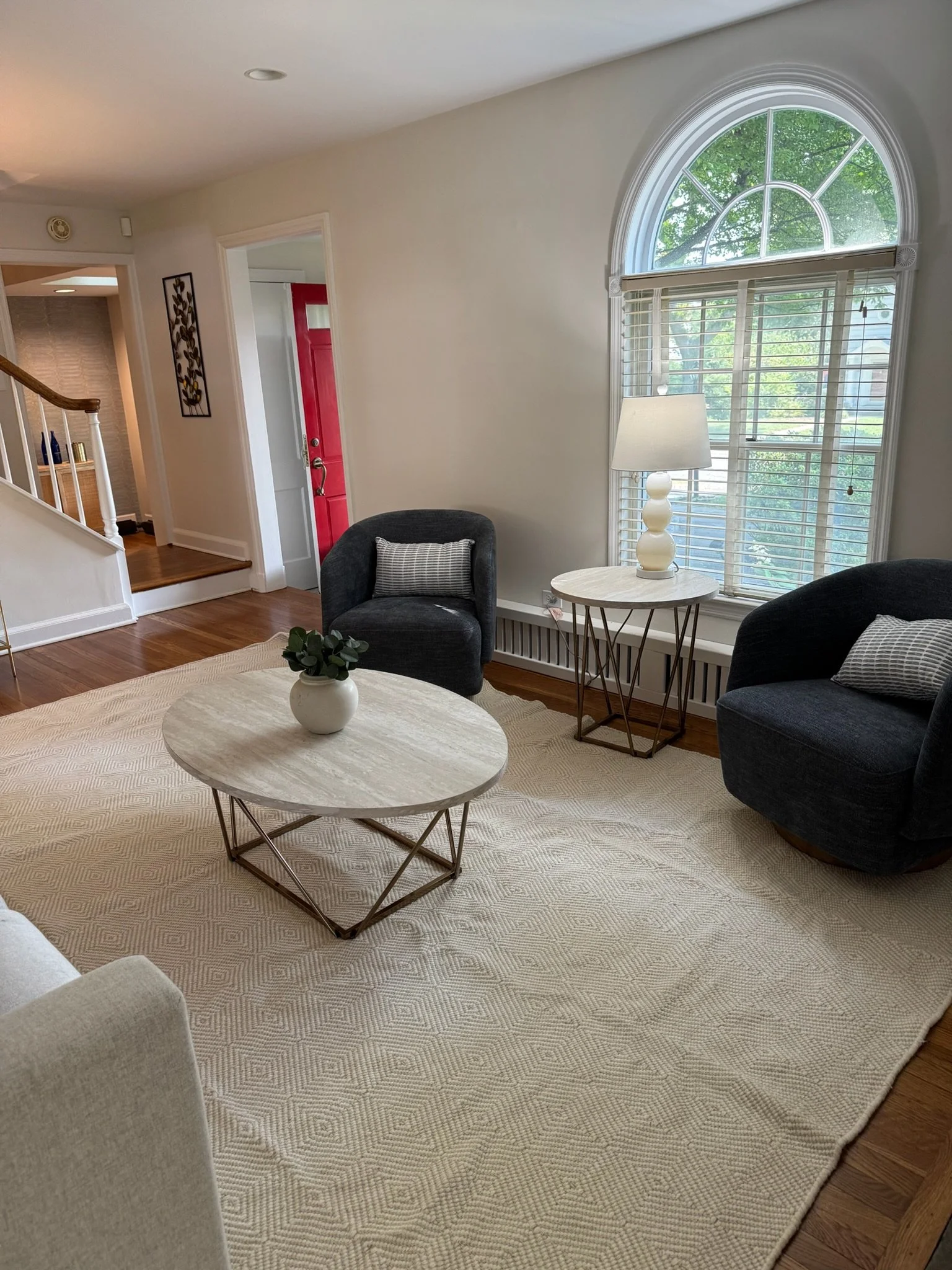 A living room with two dark gray armchairs, a coffee table with a potted plant, a side table with a lamp, a large window with blinds, and a cream rug on wooden flooring.