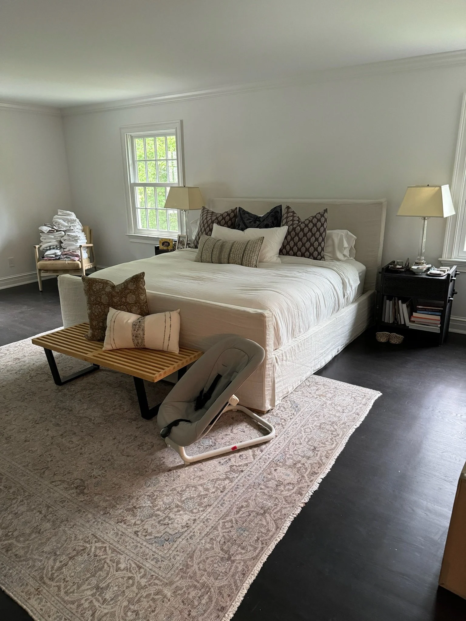 A bedroom with a white bed, several pillows, two nightstands with lamps, a window, a rug, a rocking chair, and a stack of folded clothes in the corner.