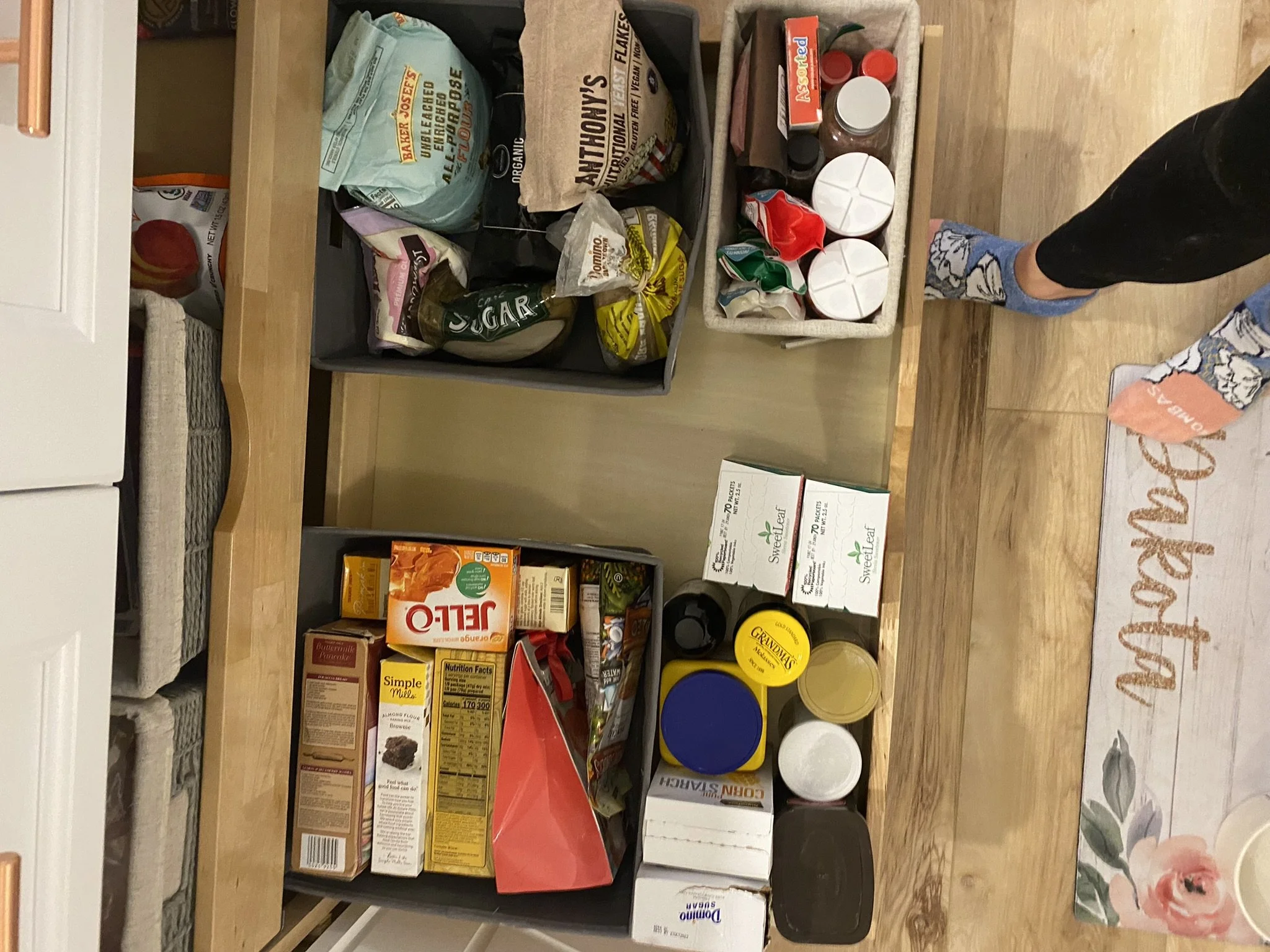 Pantry organized with various food items, canned goods, snacks, and condiments on open shelves. A small box of SweetLeaf stevia, canned and boxed foods, and plastic containers are also visible.