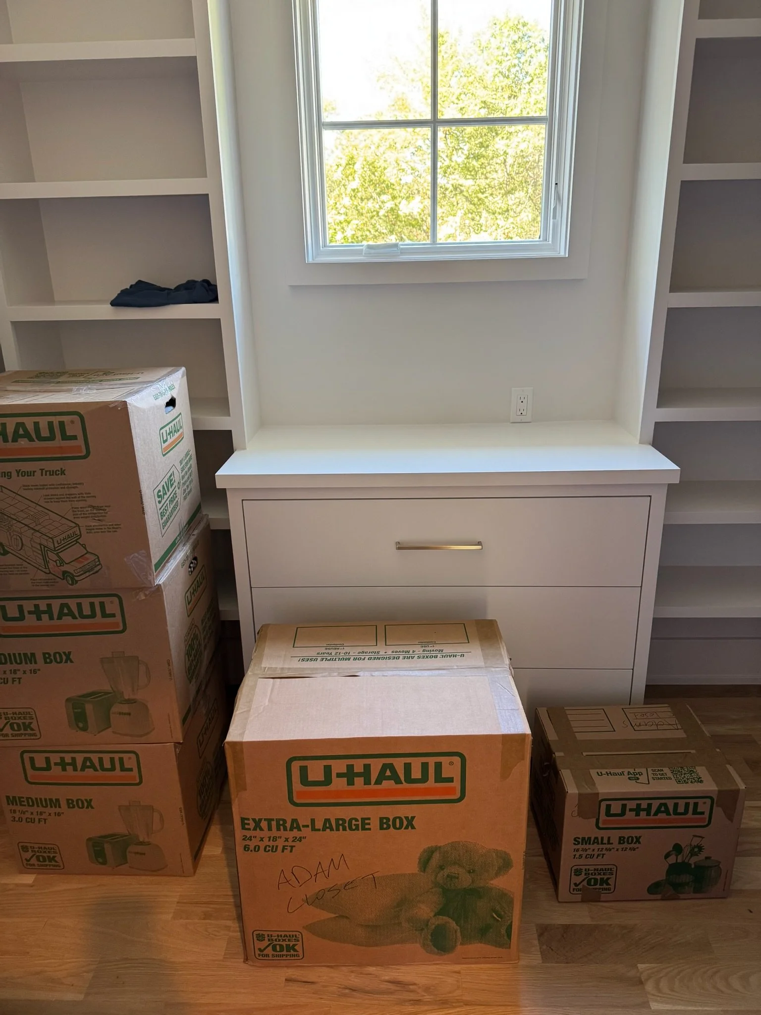 A room with a window showing trees outside, white built-in shelves on both sides, a white drawer unit beneath the window, and U-Haul boxes on the floor labeled extra-large, medium, and small.