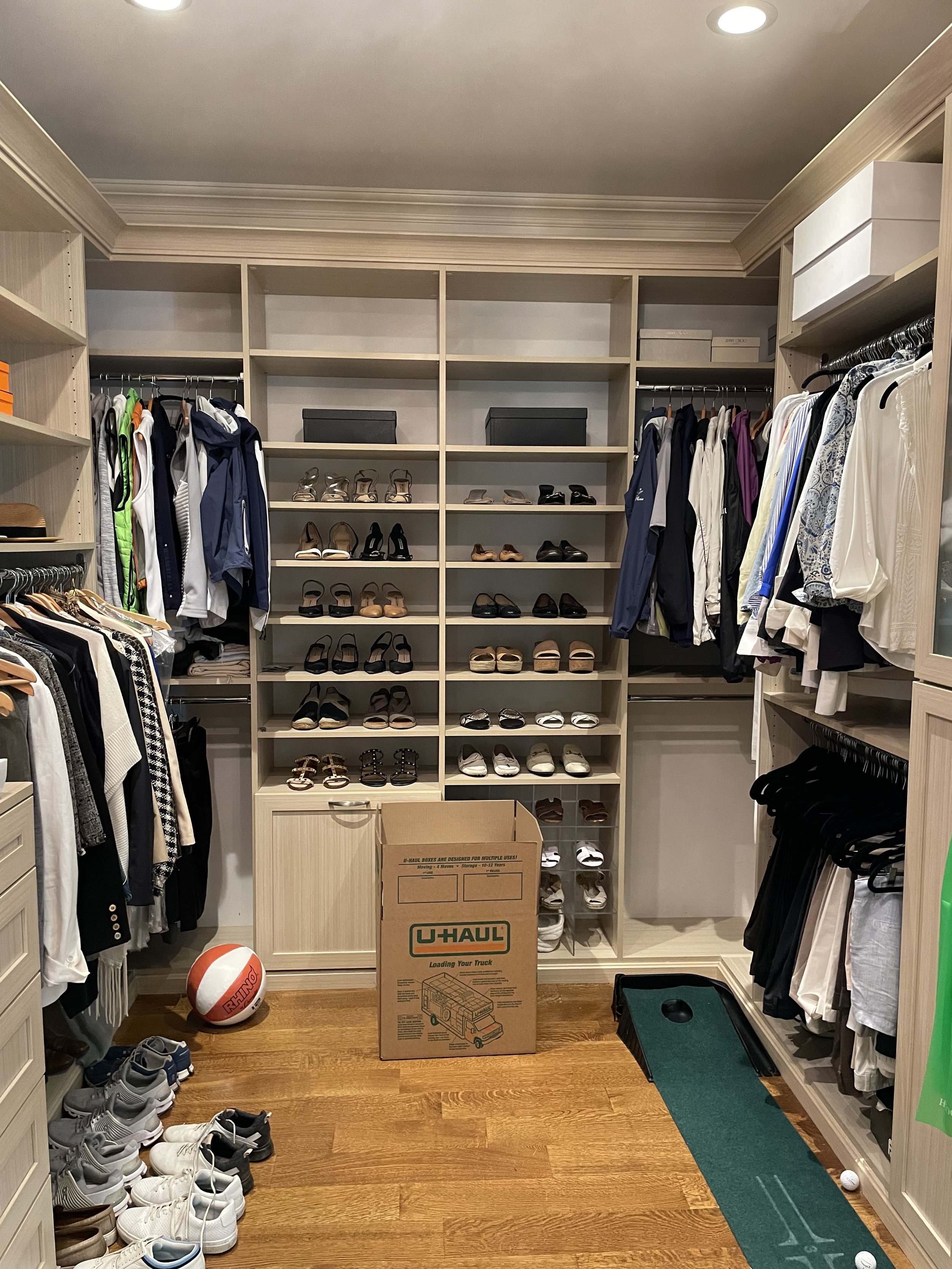 A walk-in closet with built-in shelves filled with shoes, clothing, and boxes. There are sneakers and sandals on the shelves, hanging clothes on both sides, and a U-Haul moving box in the center. The floor is wooden and there is a small golf putting mat with a golf ball and hole in the right corner.