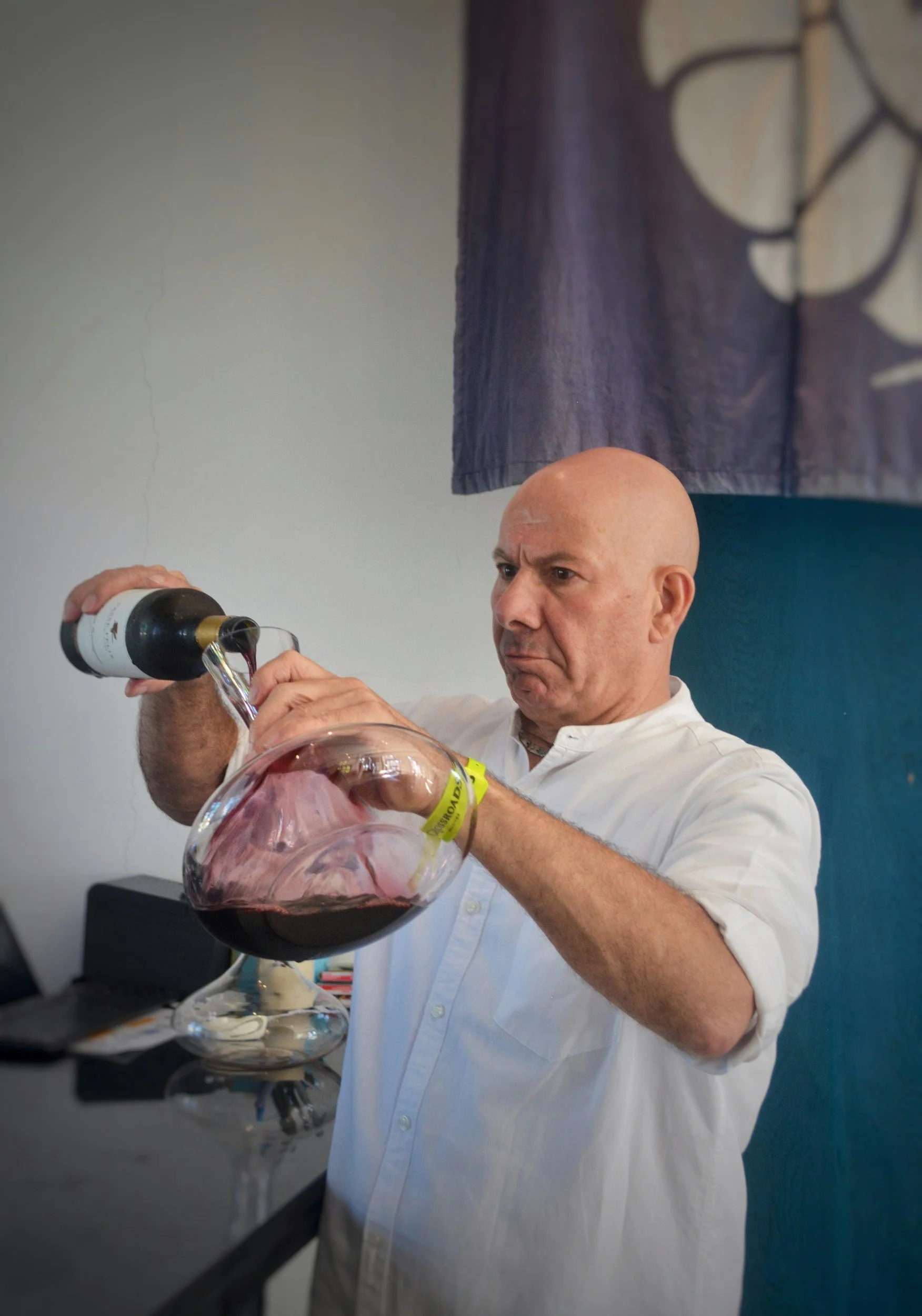 A man with a serious expression pours red wine from a bottle into a glass decanter at a bar or counter.