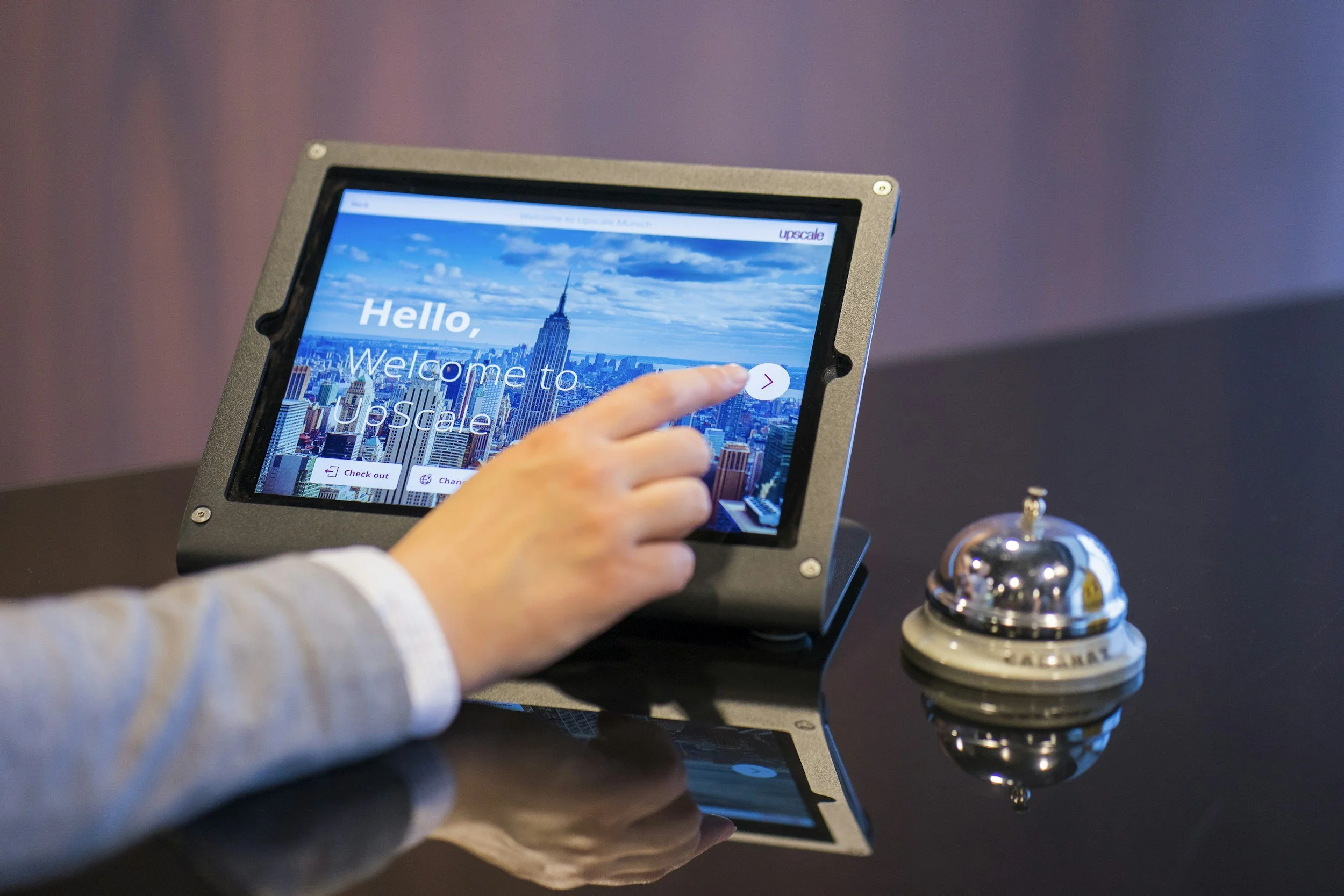 A person's hand on a touchscreen tablet displaying a welcome message for Upscale with an image of the New York City skyline and the Empire State Building in the background. A silver service bell is placed on the table beside the tablet.