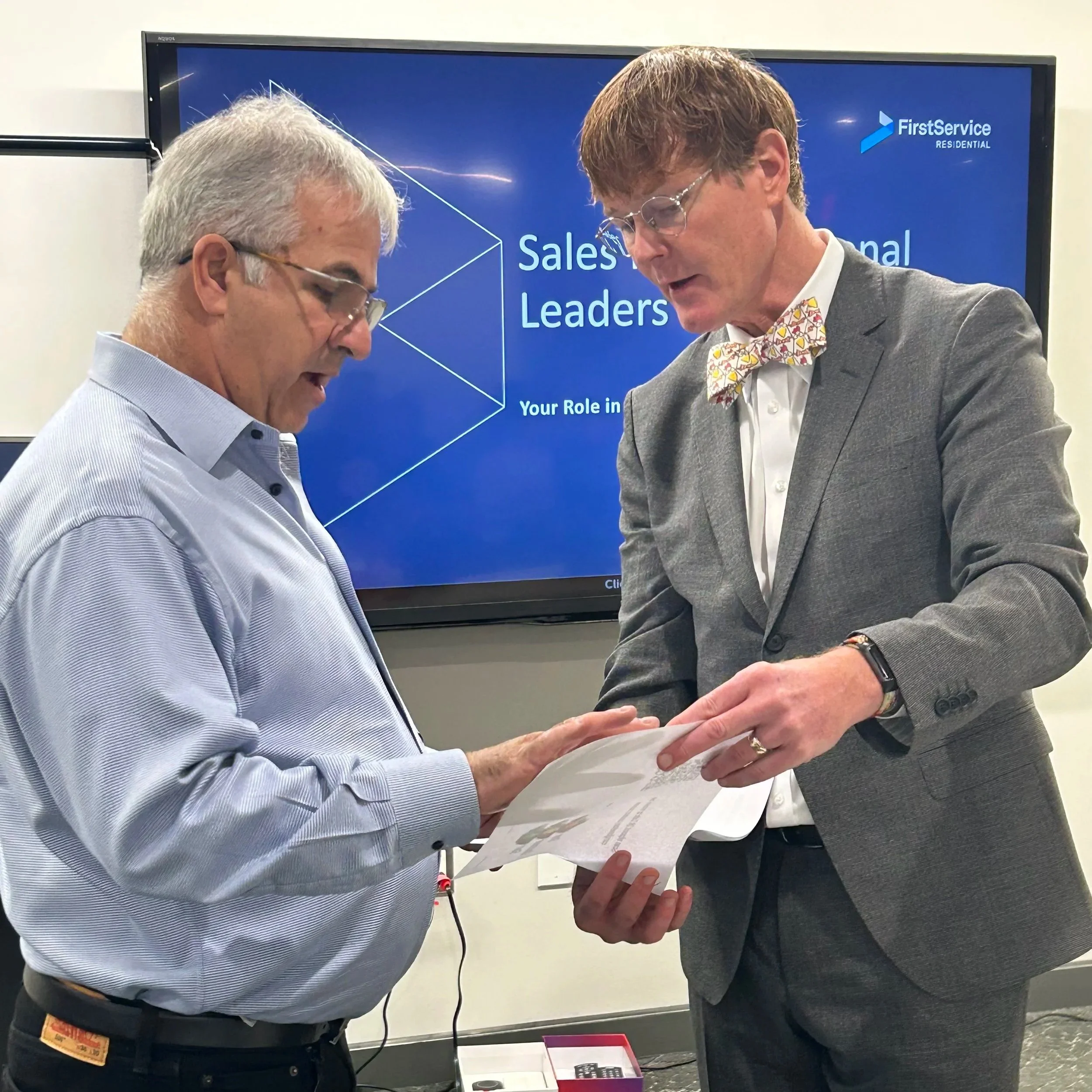 Two men in business attire looking at documents together in front of a presentation screen that reads 'Sales & Leadership' and 'Your Role in'.