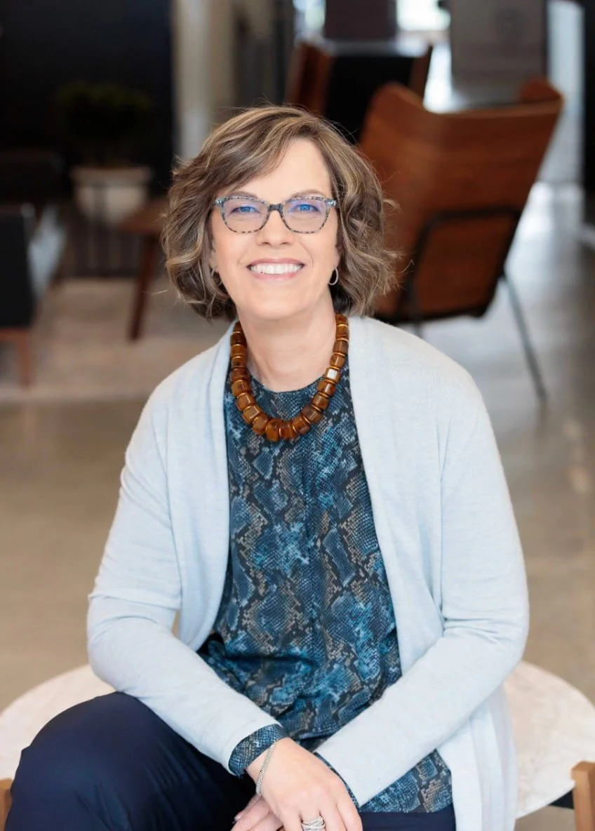 A smiling woman with short wavy hair, wearing glasses, a patterned blouse, a light cardigan, a chunky brown necklace, and sitting indoors with a wooden chair and a potted plant in the background.
