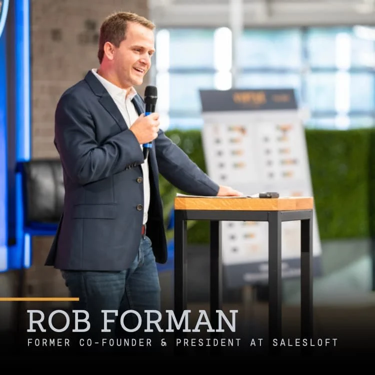 Rob Forman speaking into a microphone at a presentation or event, standing behind a small podium, in a modern indoor setting.