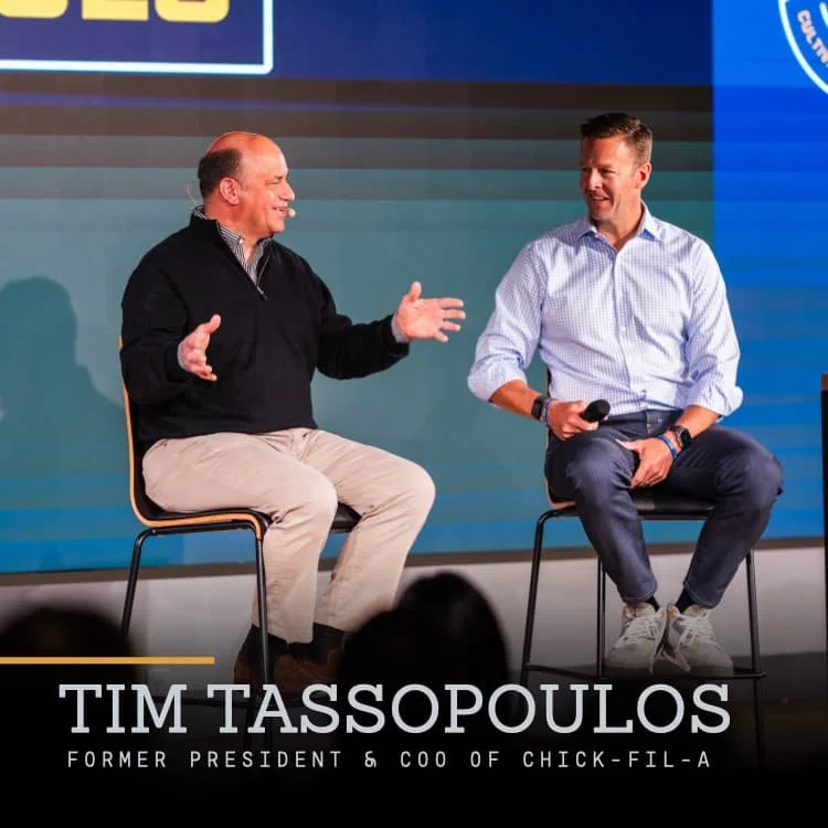 Two men sitting on stage, engaged in conversation. One is holding a microphone. The backdrop has logos and text, indicating a formal event or interview. Caption reads "Tim Tassopoulos, Former President & COO of Chick-fil-A."