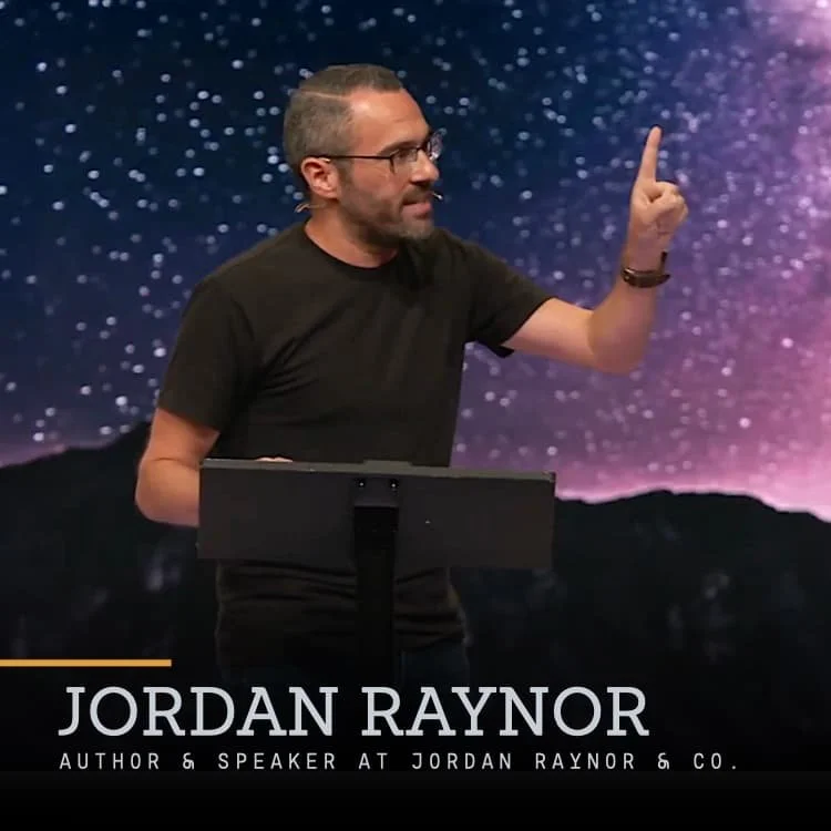 Jordan Raynor, author and speaker, standing at a podium with a backdrop of a starry night sky, gesturing upward with his right index finger.