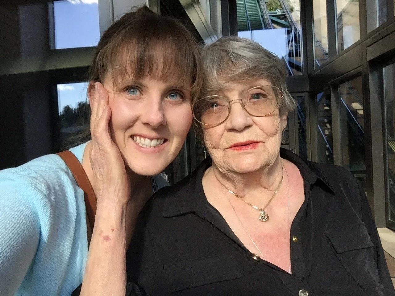 Two white women smiling at the camera for a selfie style photo. The woman on the left is young with brown bangs and blue eyes. The woman on the right is older with short gray hair and glasses.