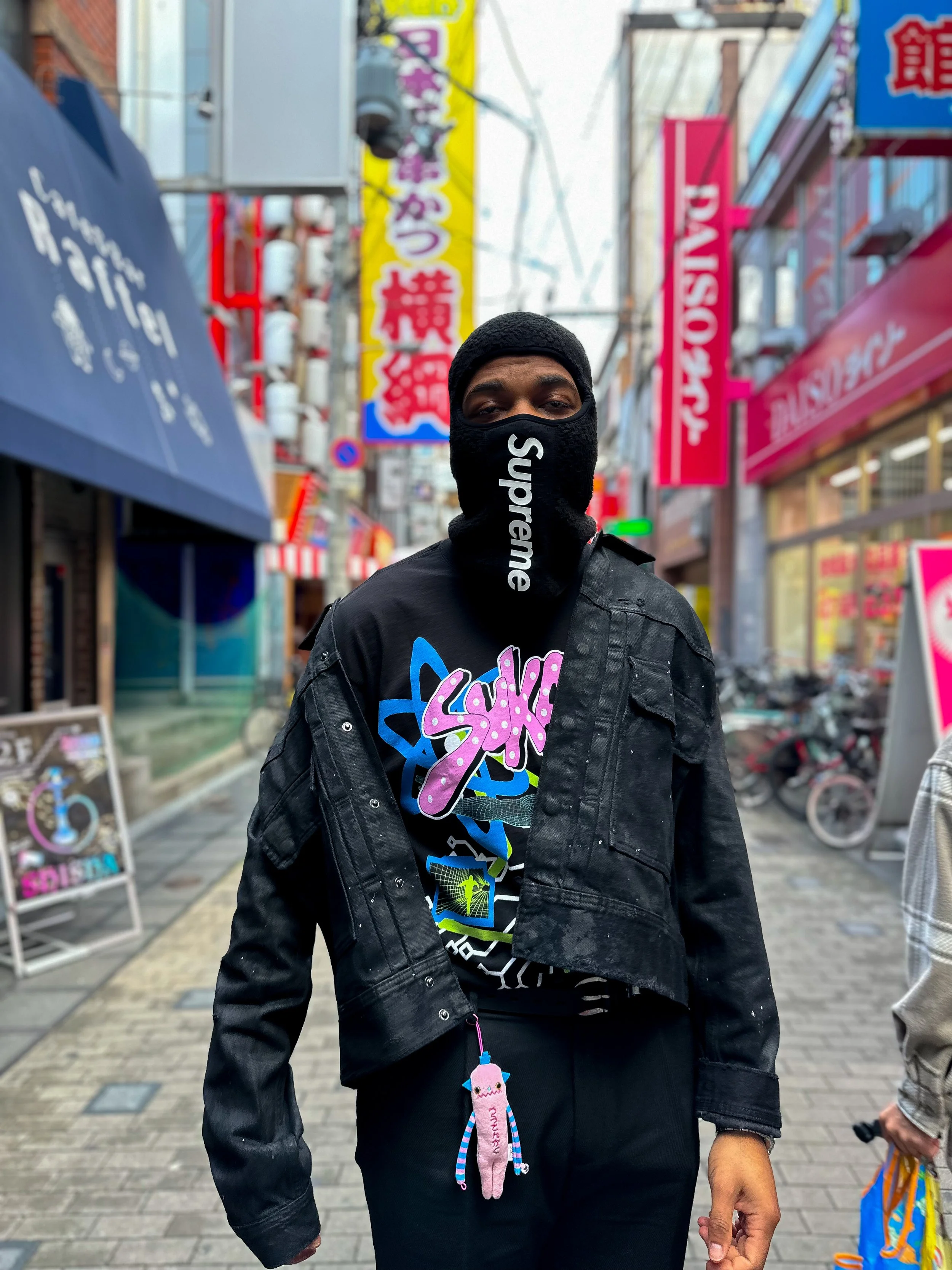 A man standing on a busy street in Japan, wearing a black Supreme mask, a black crop jacket, and a colorful graphic t-shirt, with neon signs and shops in the background.