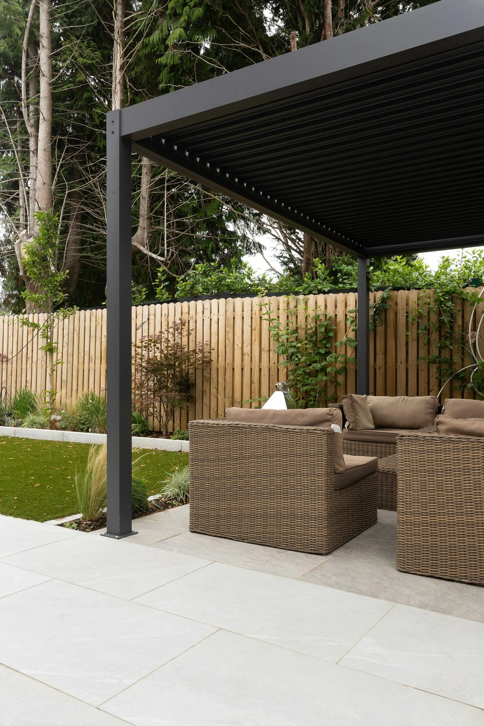 A backyard patio area with a modern black pergola, wicker outdoor sofa with beige cushions, a glass lamp, a patch of green grass, various plants, and a wooden fence with trees in the background.