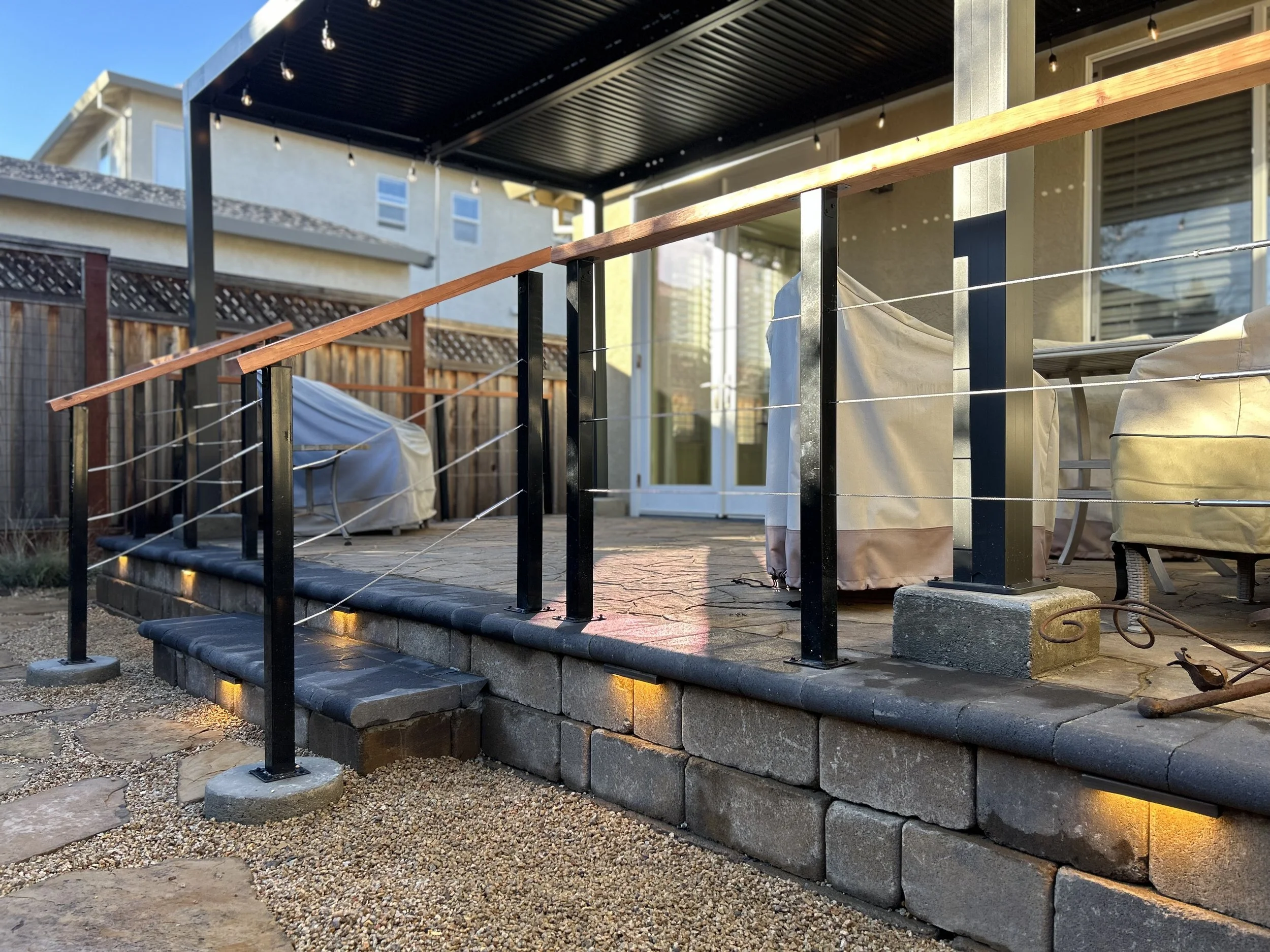 Backyard patio with a screened-in porch, stone stairs with embedded lighting, black metal and wood handrail, covered outdoor furniture, covered grill, wooden fencing, and string lights hanging from the porch ceiling.