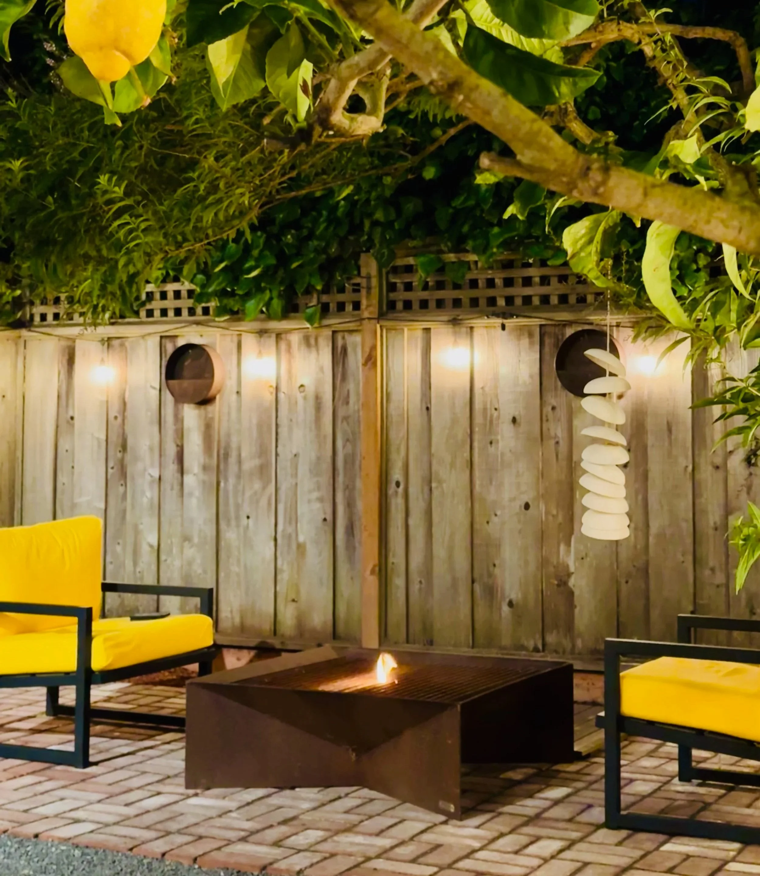 Outdoor patio with two yellow cushioned chairs around a fire pit, wooden fence with lights, hanging art piece, and leafy trees overhead.