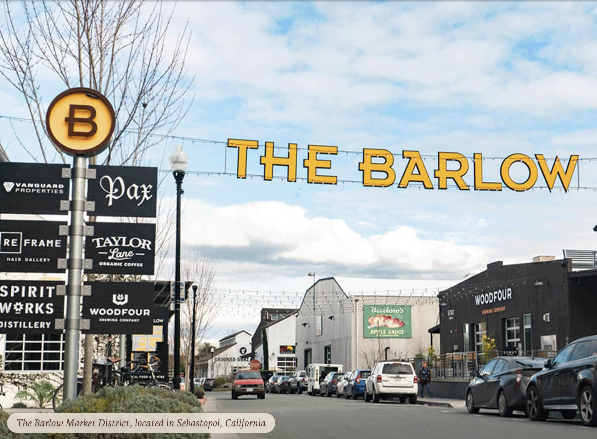 View of The Barlow Market District sign in Sebastopol, California, with shops, parked cars, and pedestrians, under a partly cloudy sky.
