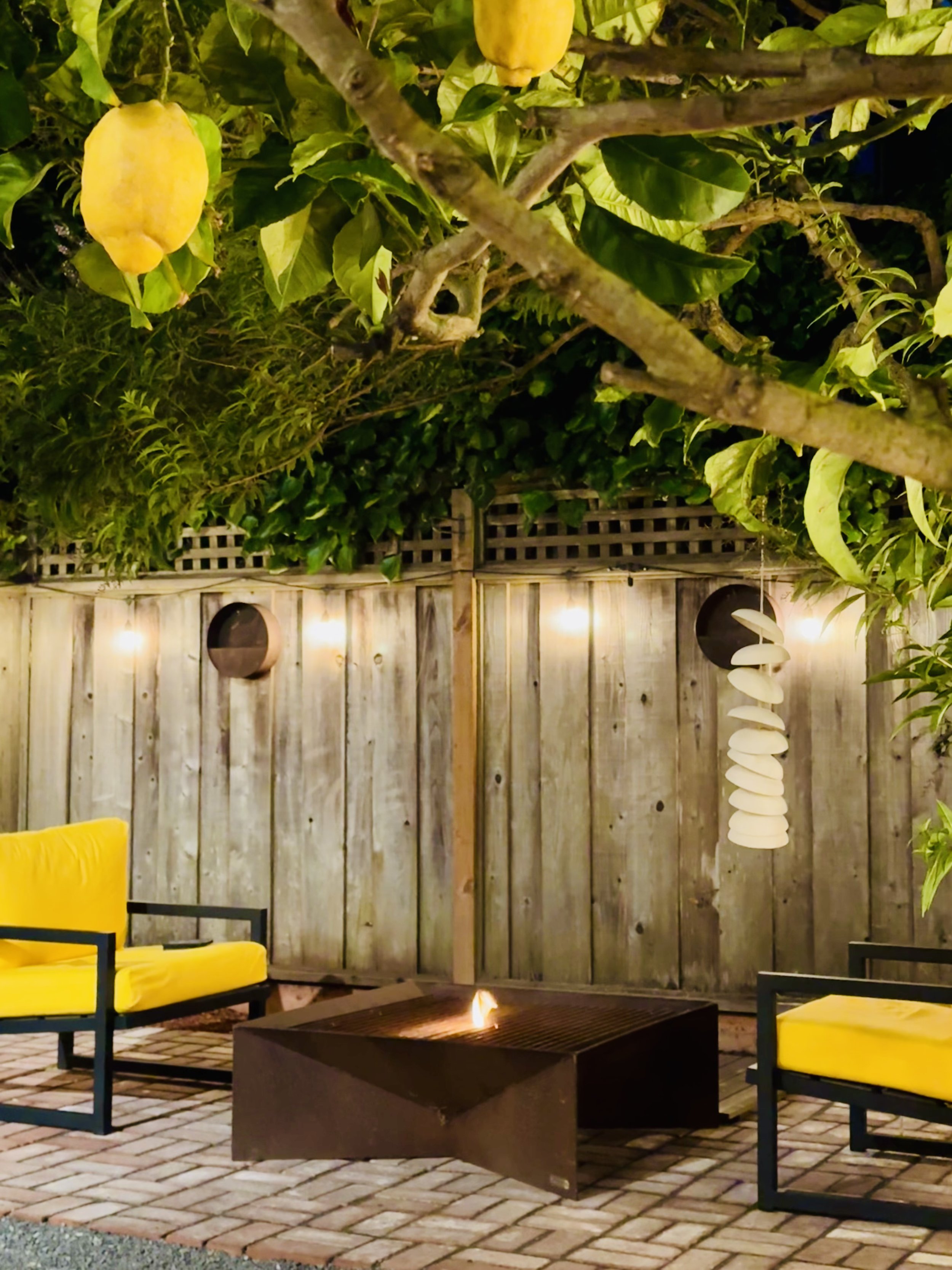 An outdoor patio area with yellow cushioned chairs, a fire pit, and a wooden privacy fence. The scene is illuminated with wall lights and decorative lighting, with trees and green foliage overhead in san francisco
