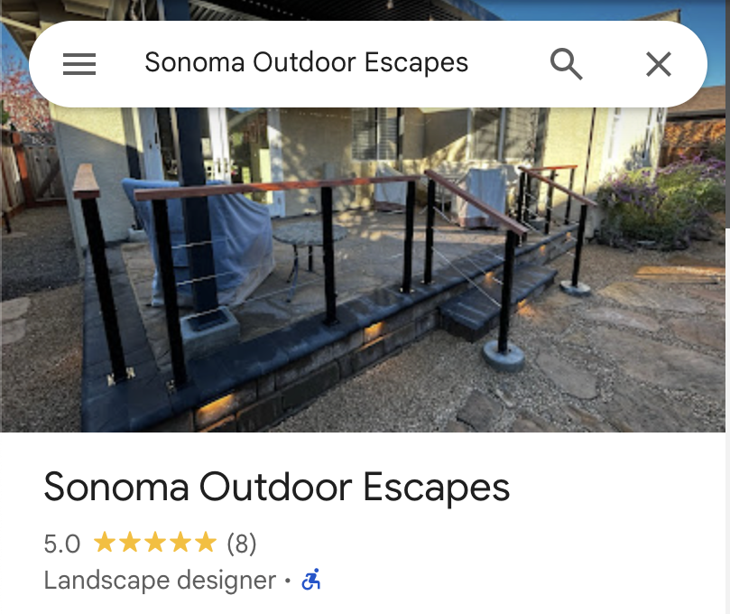 A small outdoor patio with a black brick border, concrete steps, a railing, and some furniture covered with a tarp, located in a backyard with a house and some plants in the background.