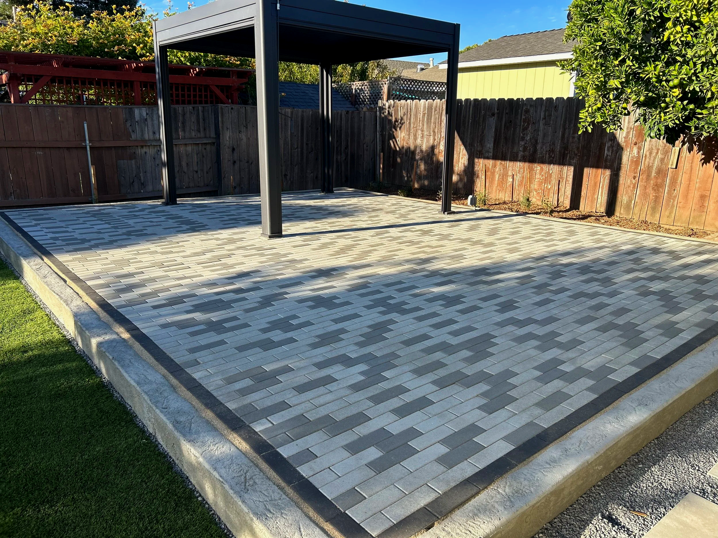 A backyard patio with gray interlocking pavers, a black metal pergola, and a wooden fence surrounding the area.