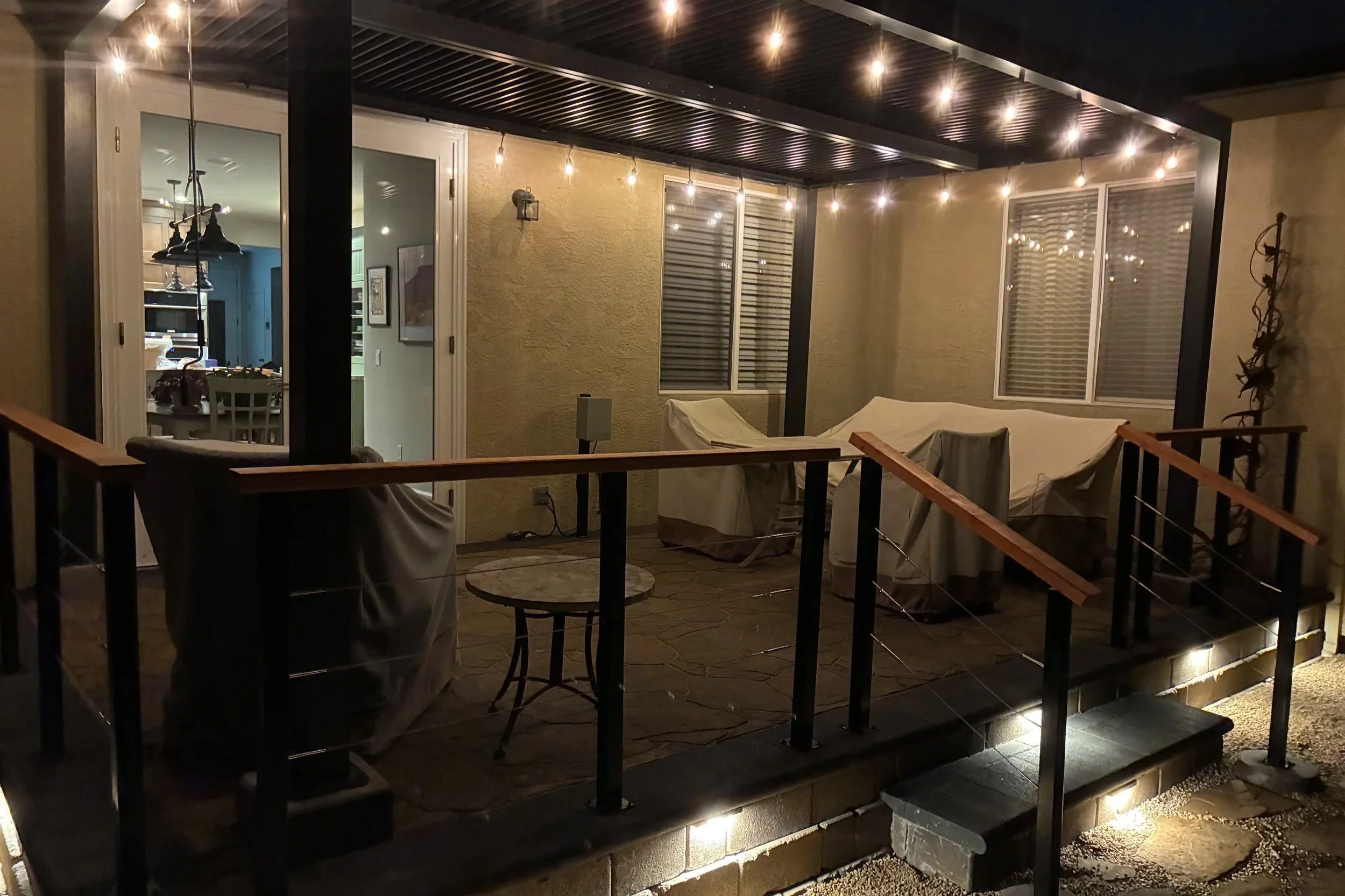 Back patio area at night with string lights hanging from the ceiling. Covered outdoor space with two covered seating areas, one with a white cloth-covered furniture and the other with a wicker chair. Through glass doors, interior of a home is visible with a dining table and chairs, and kitchen area in the background.