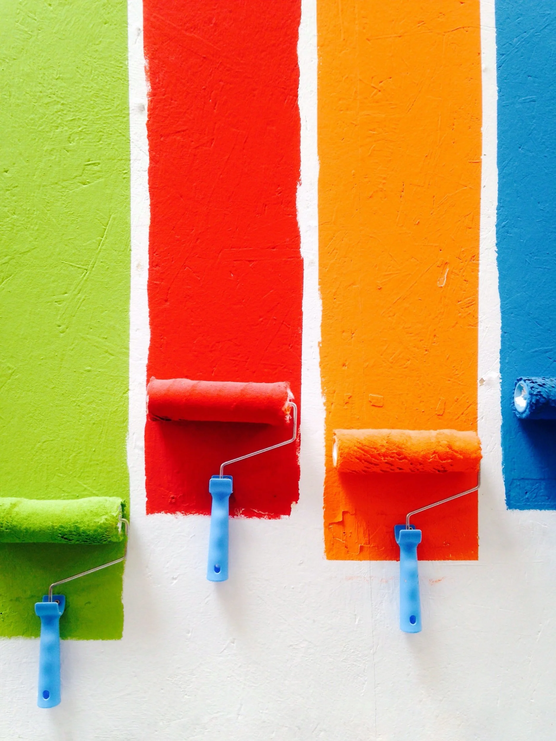Four paint rollers with blue handles applying green, red, orange, and blue paint on a white wall.