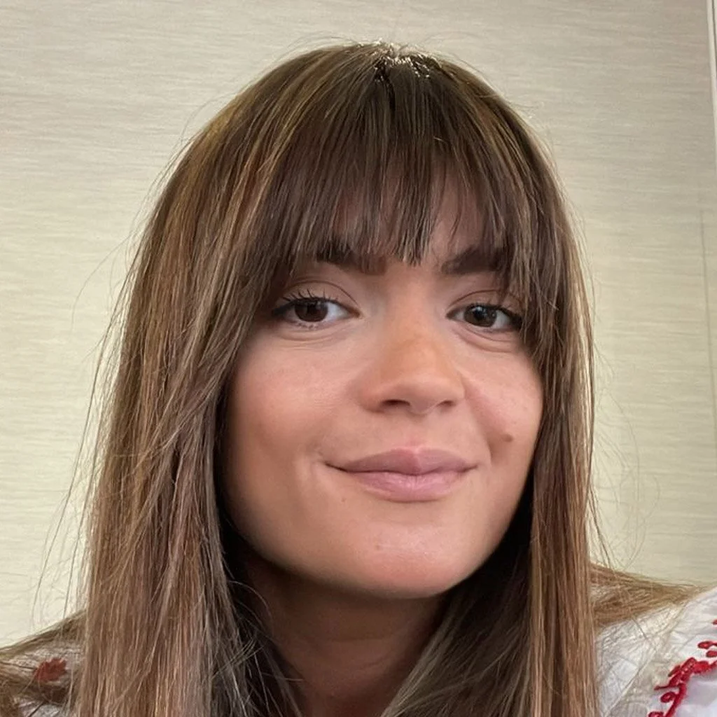 Close-up of a woman with light brown hair, straight bangs, and a subtle smile, standing against a neutral background.