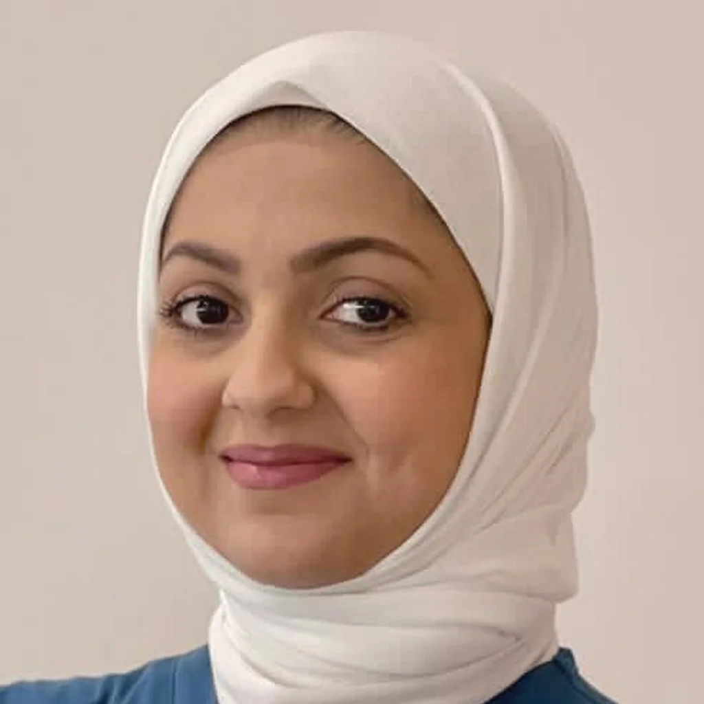 A woman with dark eyes and light makeup, wearing a white hijab and a blue top, smiling at the camera.
