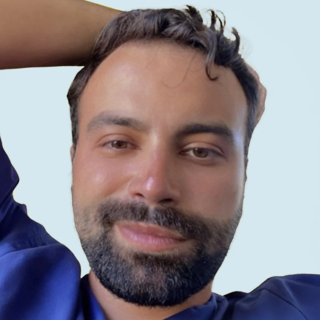 Close-up of a man with dark hair, beard, and mustache, wearing a navy blue shirt, with one hand behind his head, against a plain light background.